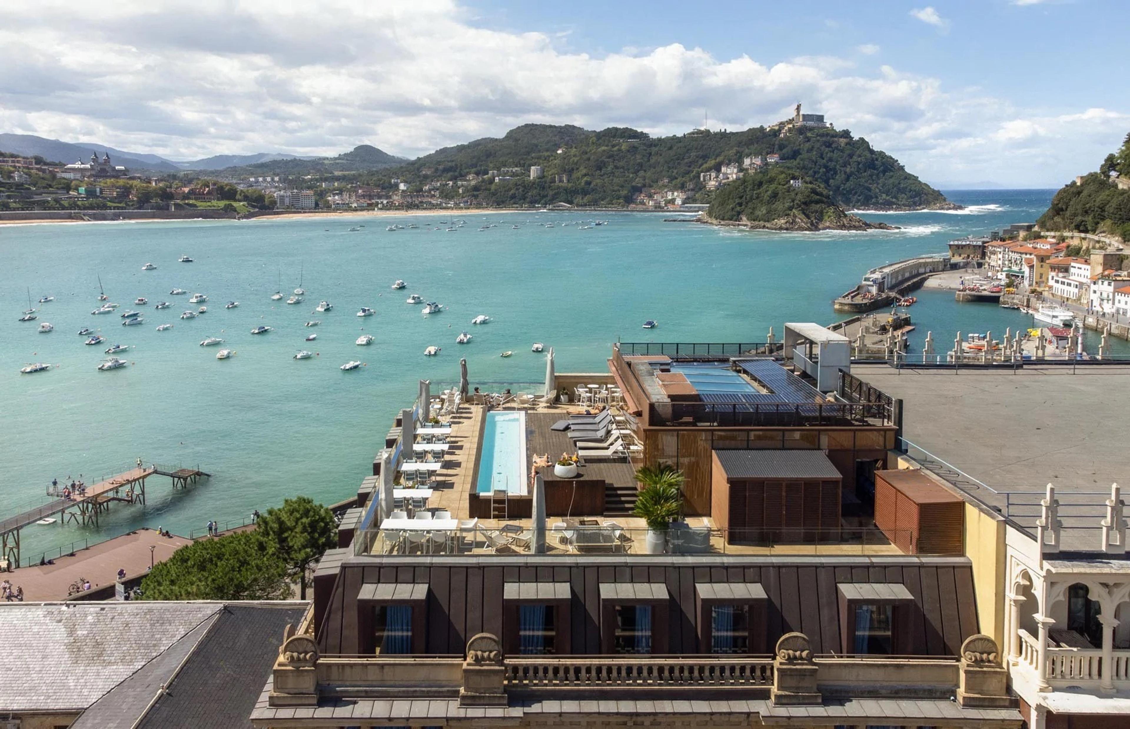A hotel building seen from above, its rooftop lined with a swimming pool, sun loungers and umbrellas, beside a wide bay with a sandy beach, moored boats and a hill topped with a building.