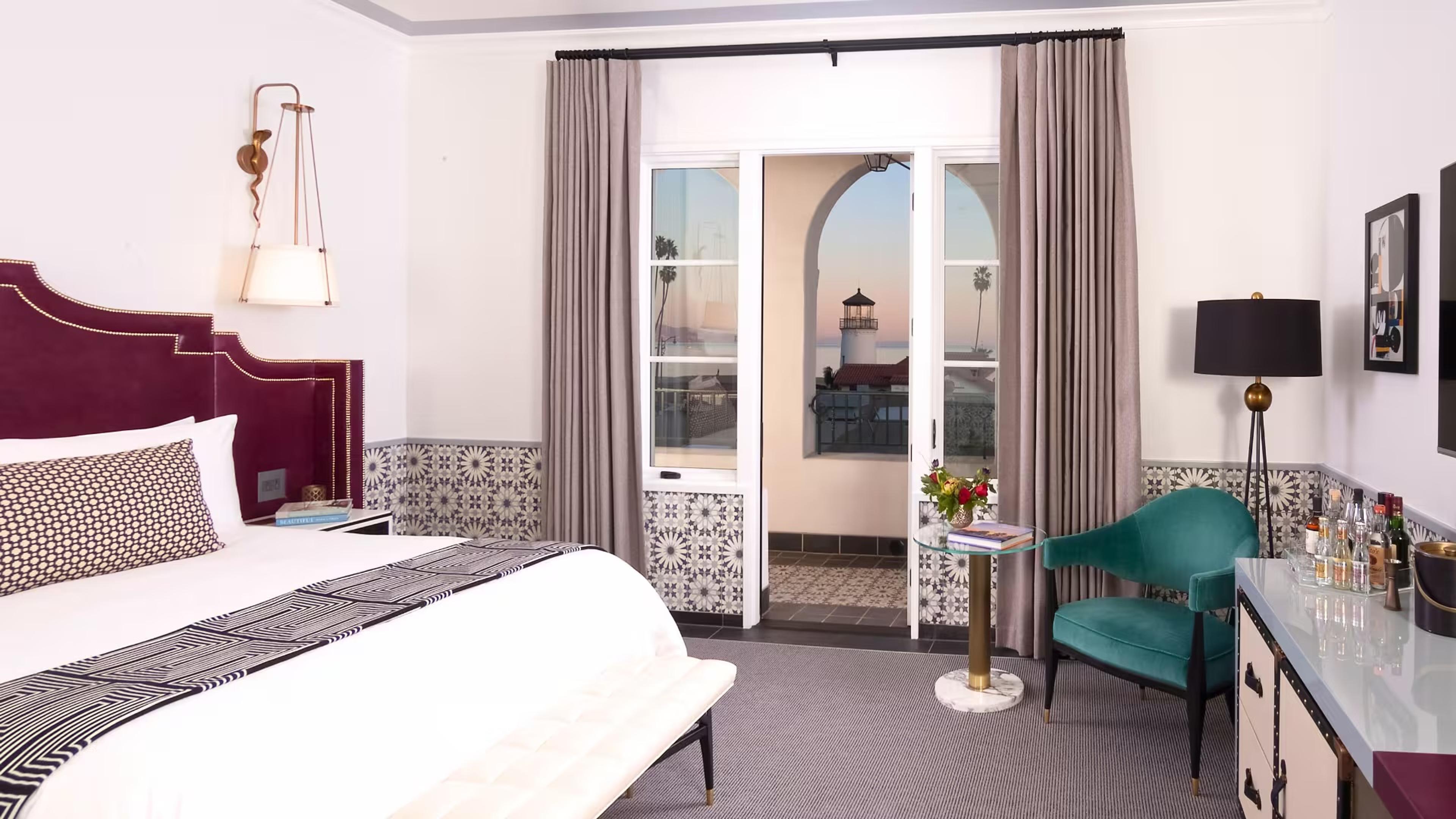 A bedroom with a maroon leather headboard and patterned throw, facing open French doors to a balcony with a view of a lighthouse and the ocean at dusk.
