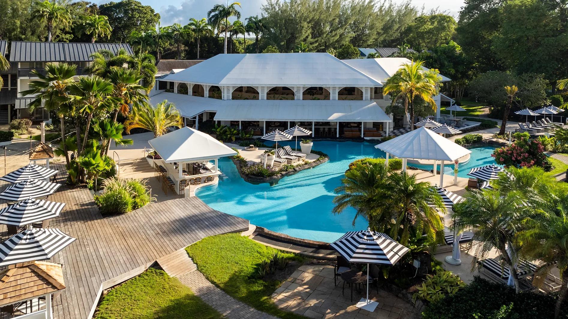 The white colonial main house seen from above, its pool curving through lawns, palms and striped parasols