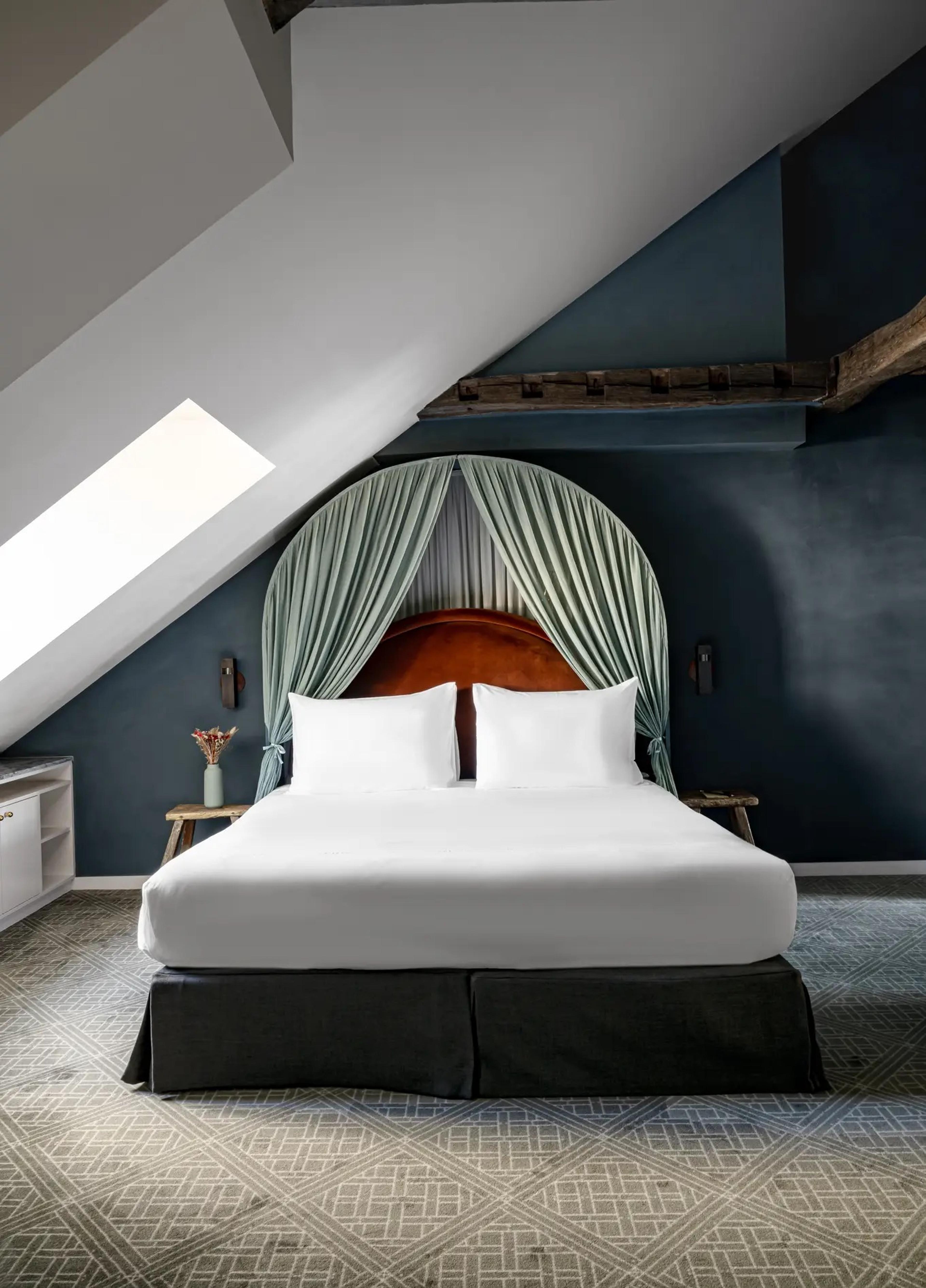 A bed with white linen and a curved orange headboard sits beneath a sage green draped canopy, in a dark blue attic room with an exposed wooden beam.