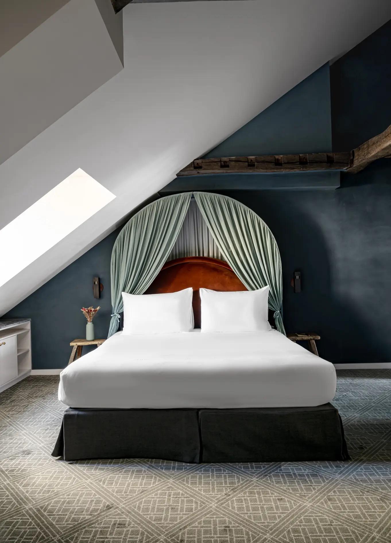 A bed with white linen and a curved orange headboard sits beneath a sage green draped canopy, in a dark blue attic room with an exposed wooden beam.