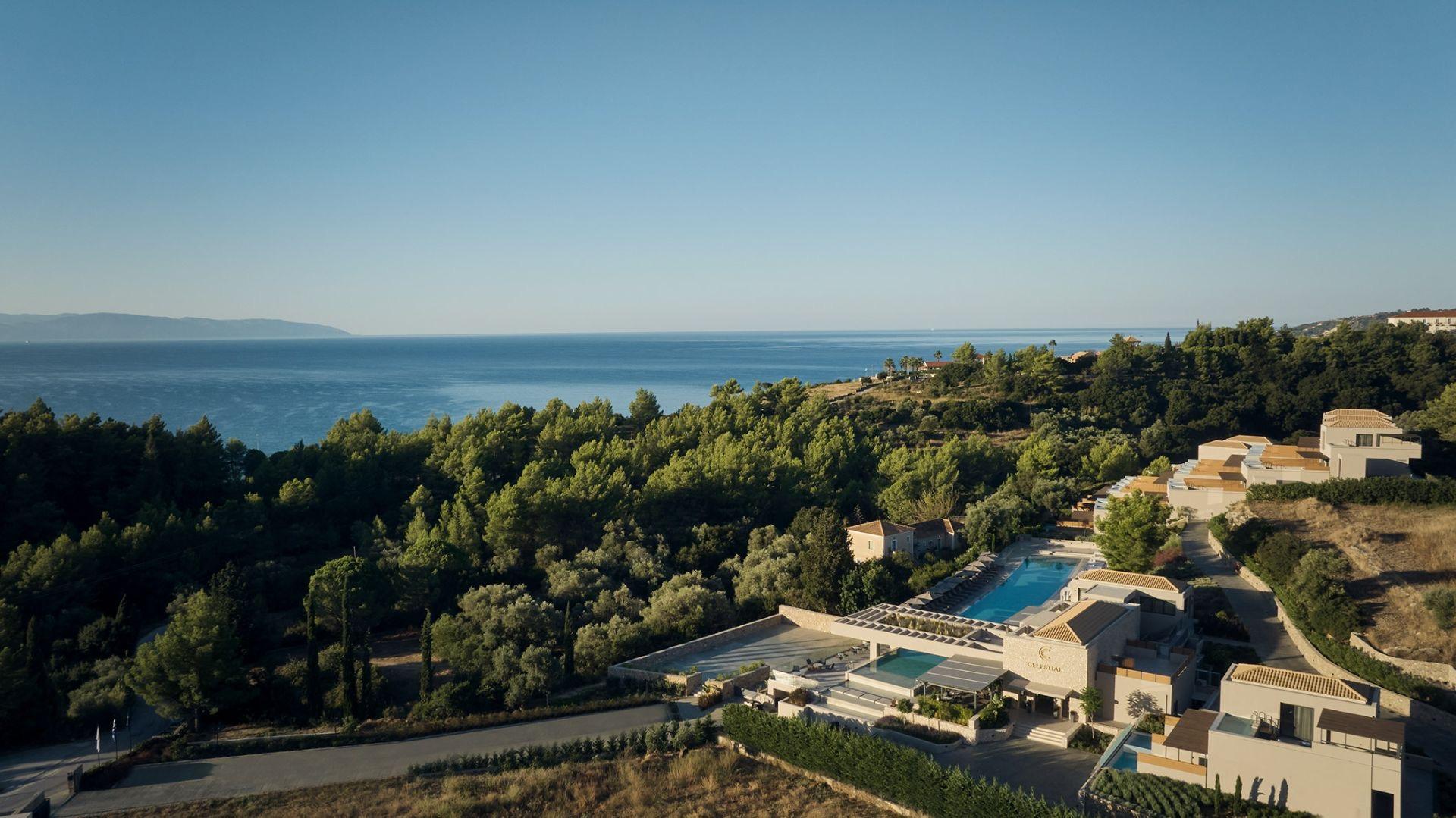 An aerial view of a hotel on a wooded hillside above the sea, with a pool and cluster of buildings in the foreground and the coastline stretching into the distance.