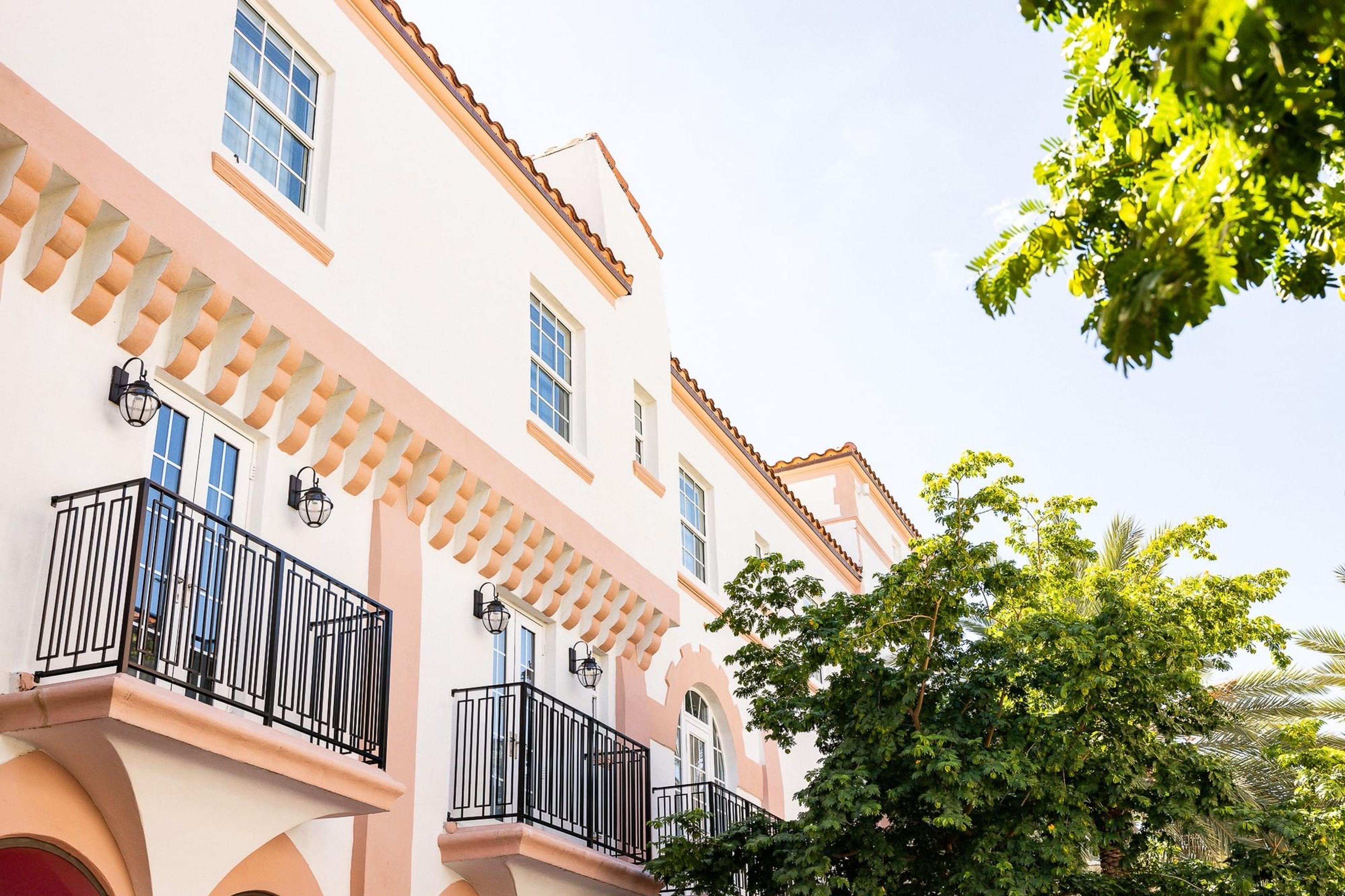 Looking up at a cream and pink Mediterranean-revival hotel facade with wrought iron balconies, arched window surrounds, and a tree in leaf at the edge of the frame.
