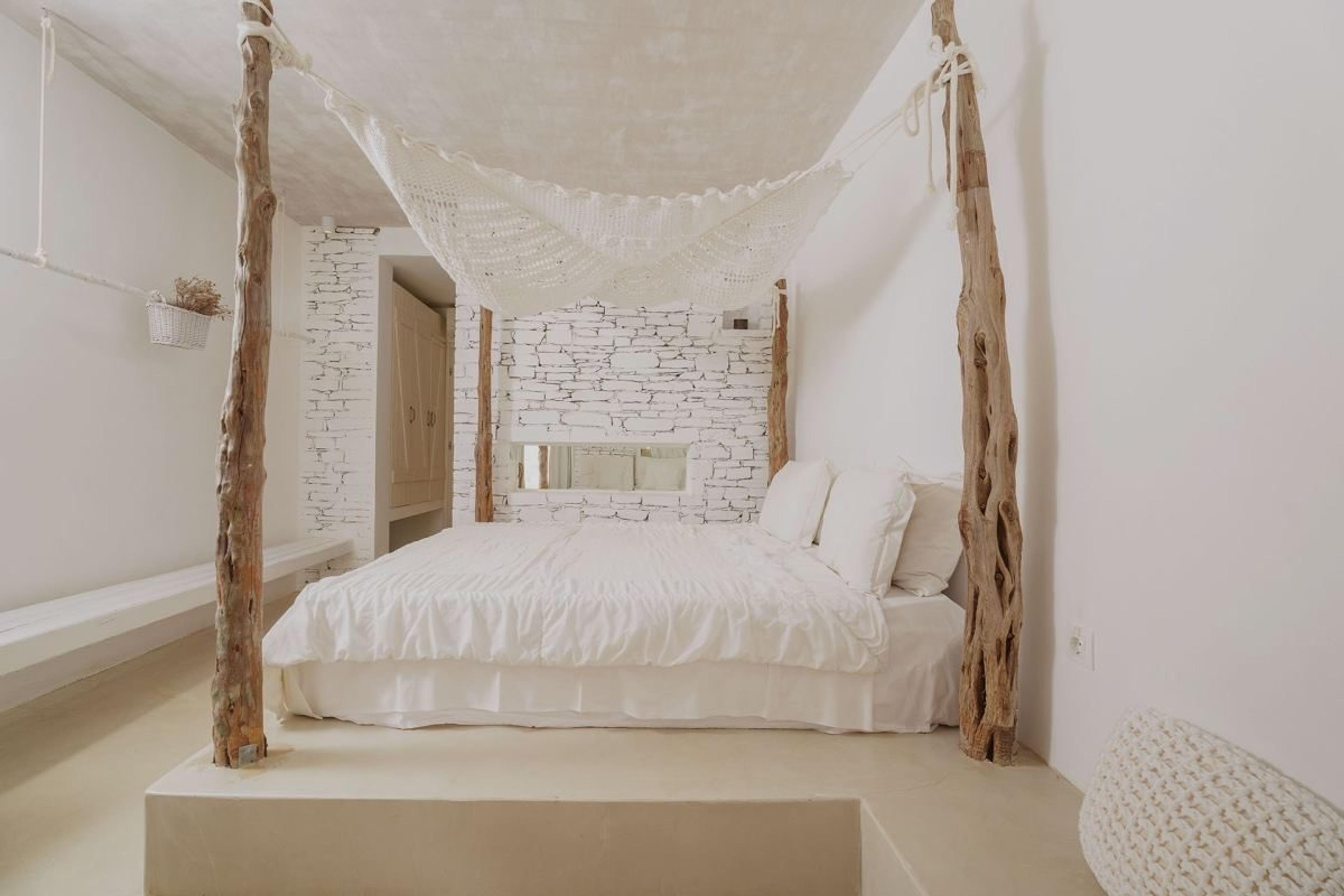 A bed on a raised platform with a driftwood post at each corner and a rope net above it, against a stone wall.