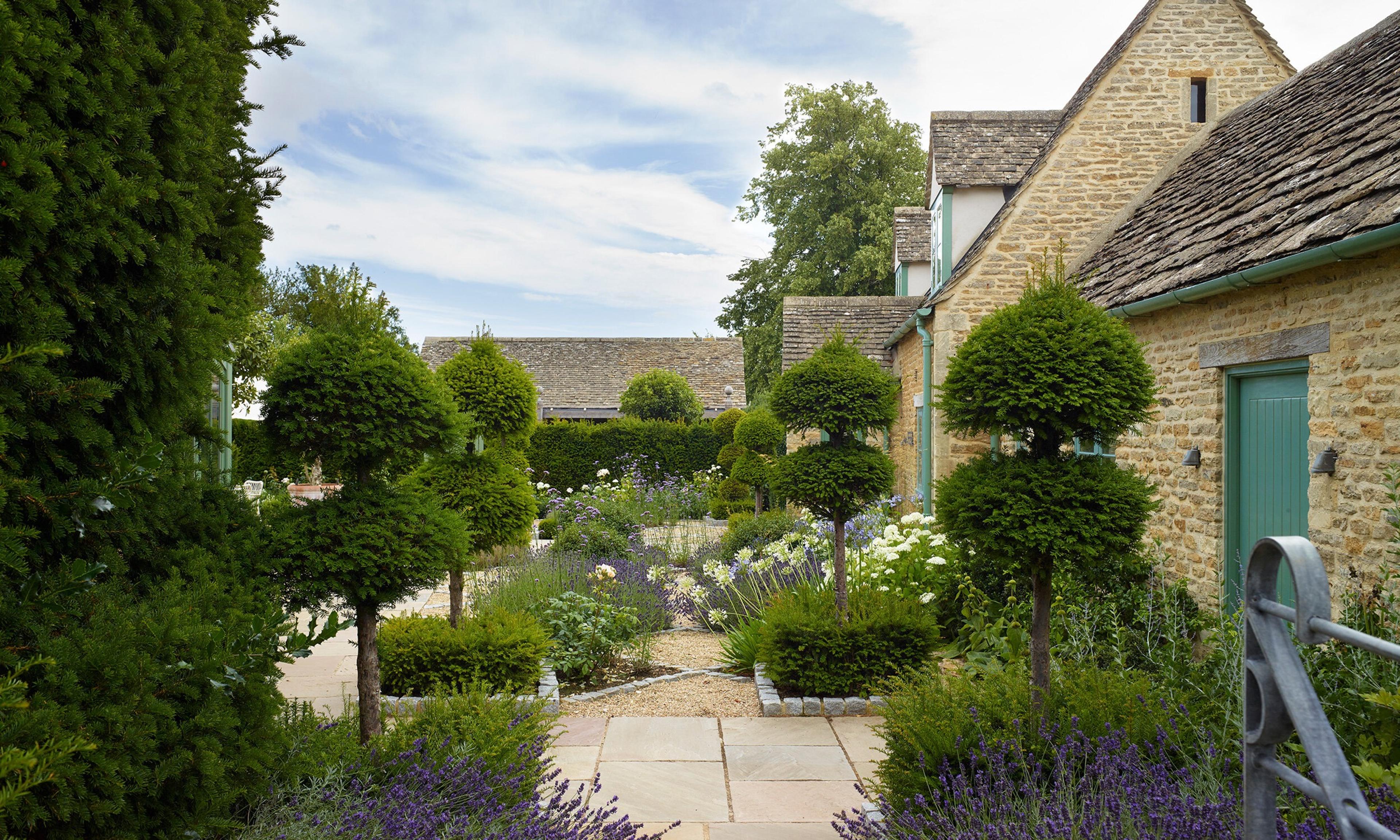 A gravel and stone path through a walled garden lined with clipped topiary and lavender, leading toward a row of stone cottages.