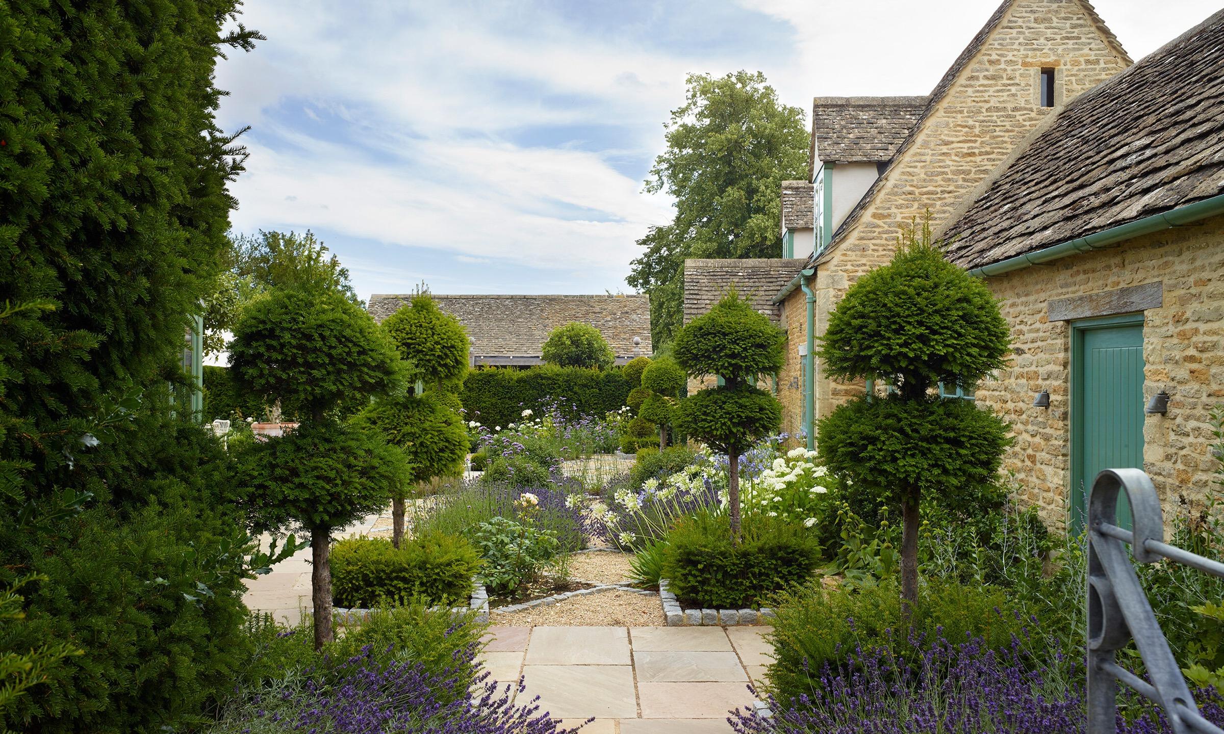 A gravel and stone path through a walled garden lined with clipped topiary and lavender, leading toward a row of stone cottages.