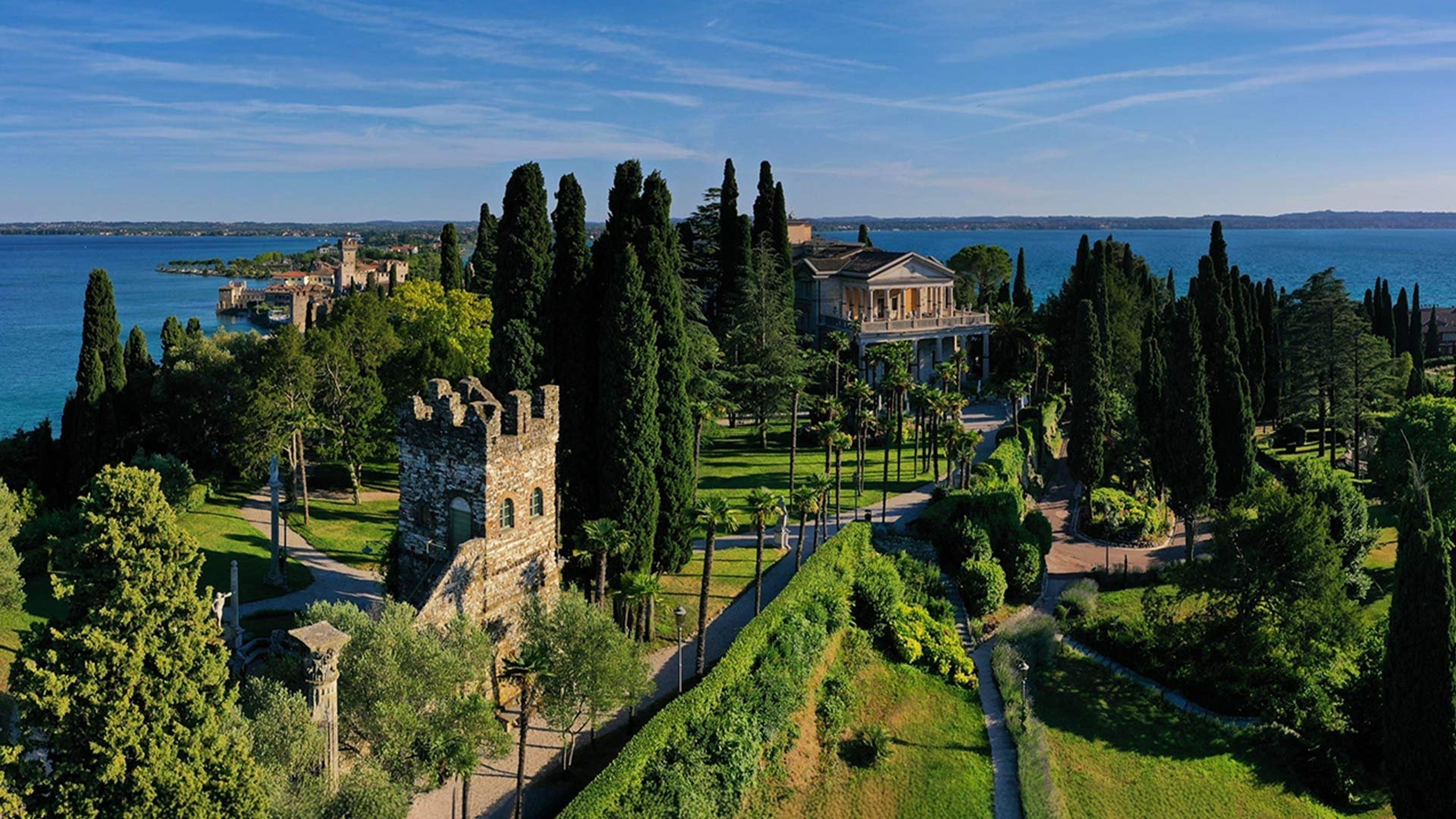 An aerial daytime view of a lakeside villa and gardens, with a stone tower, cypress trees and a walled town on the water beyond.