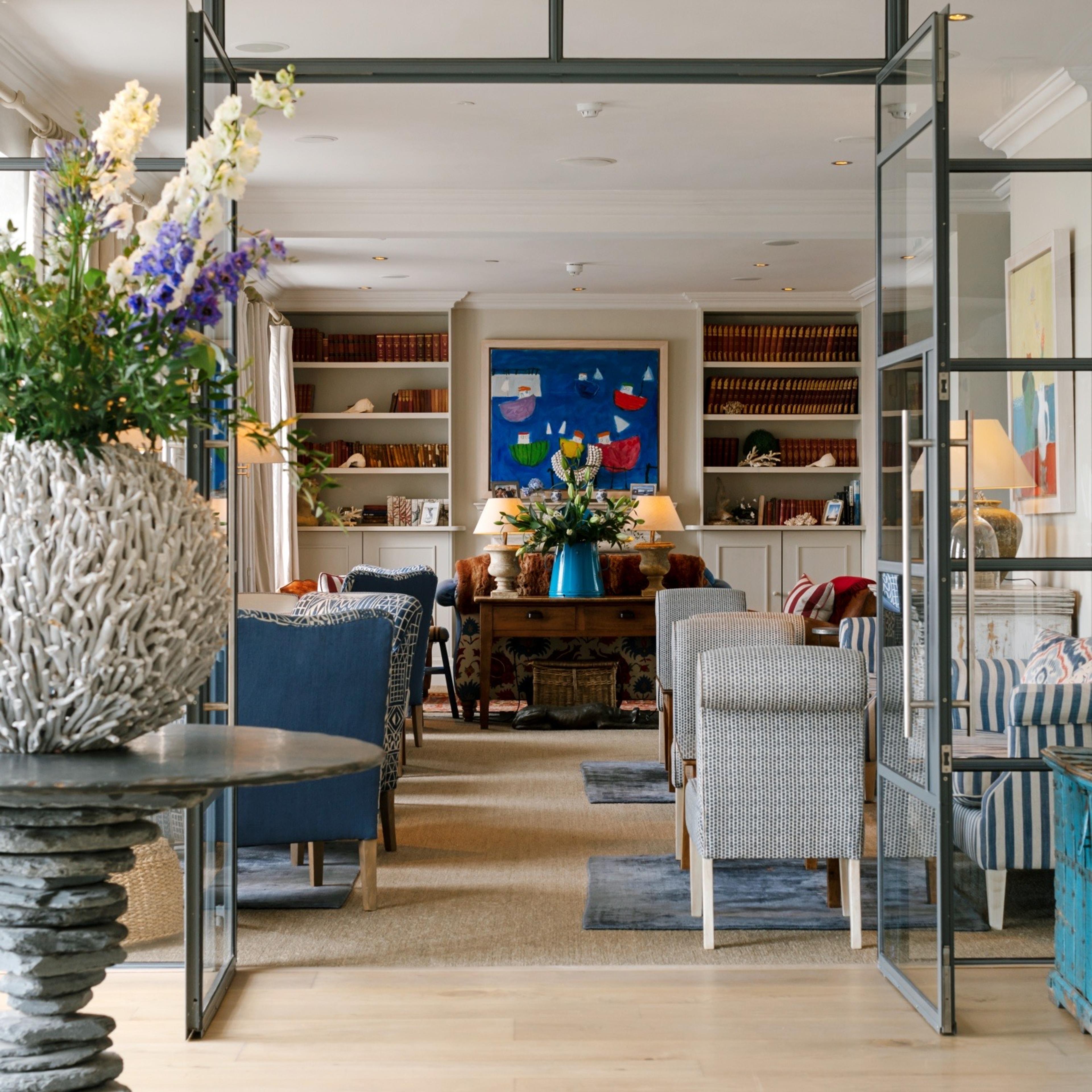 A hotel lounge with bookshelves either side of a blue painting, armchairs upholstered in blue and striped fabric, and a vase of flowers on a side table, seen through an open glazed door.