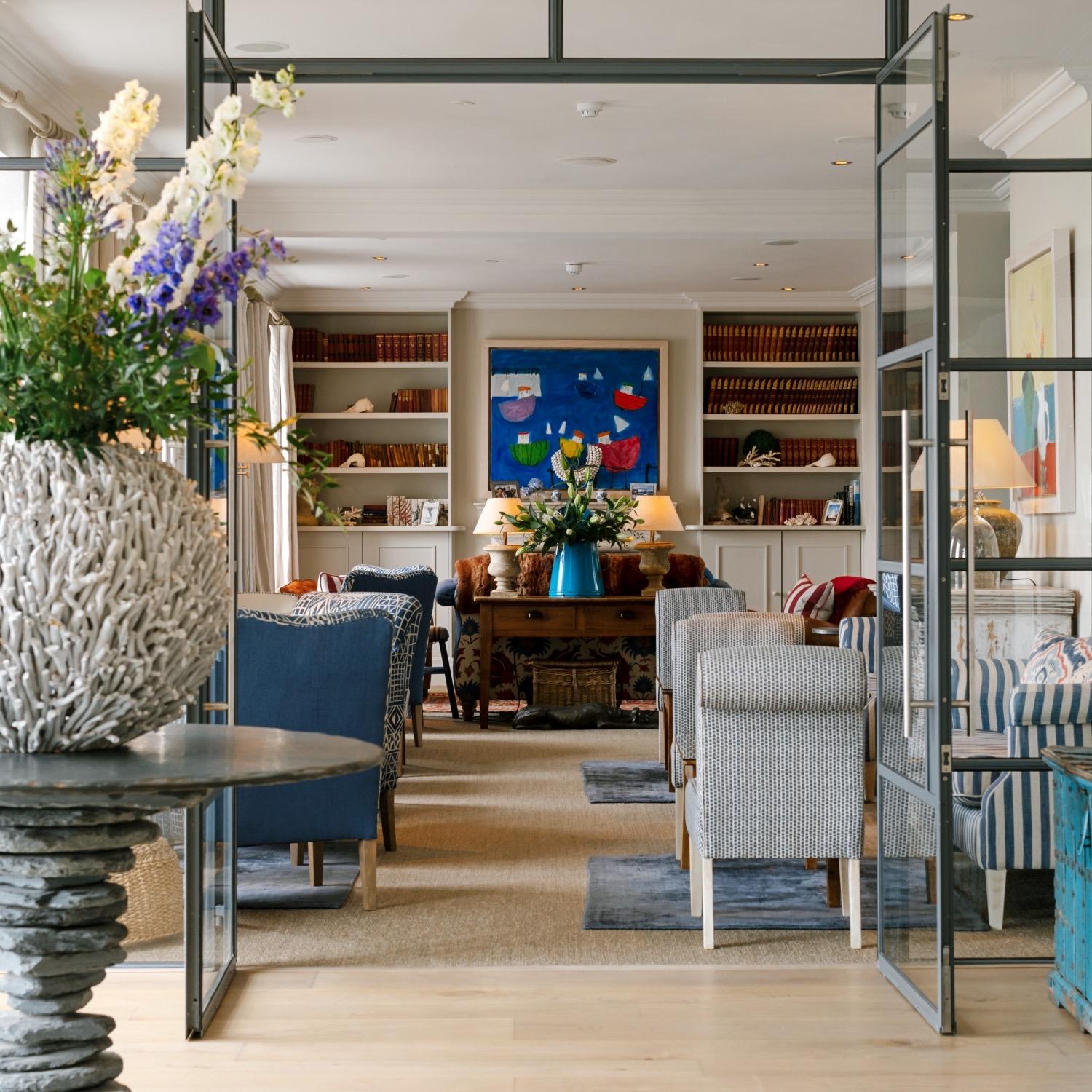 A hotel lounge with bookshelves either side of a blue painting, armchairs upholstered in blue and striped fabric, and a vase of flowers on a side table, seen through an open glazed door.