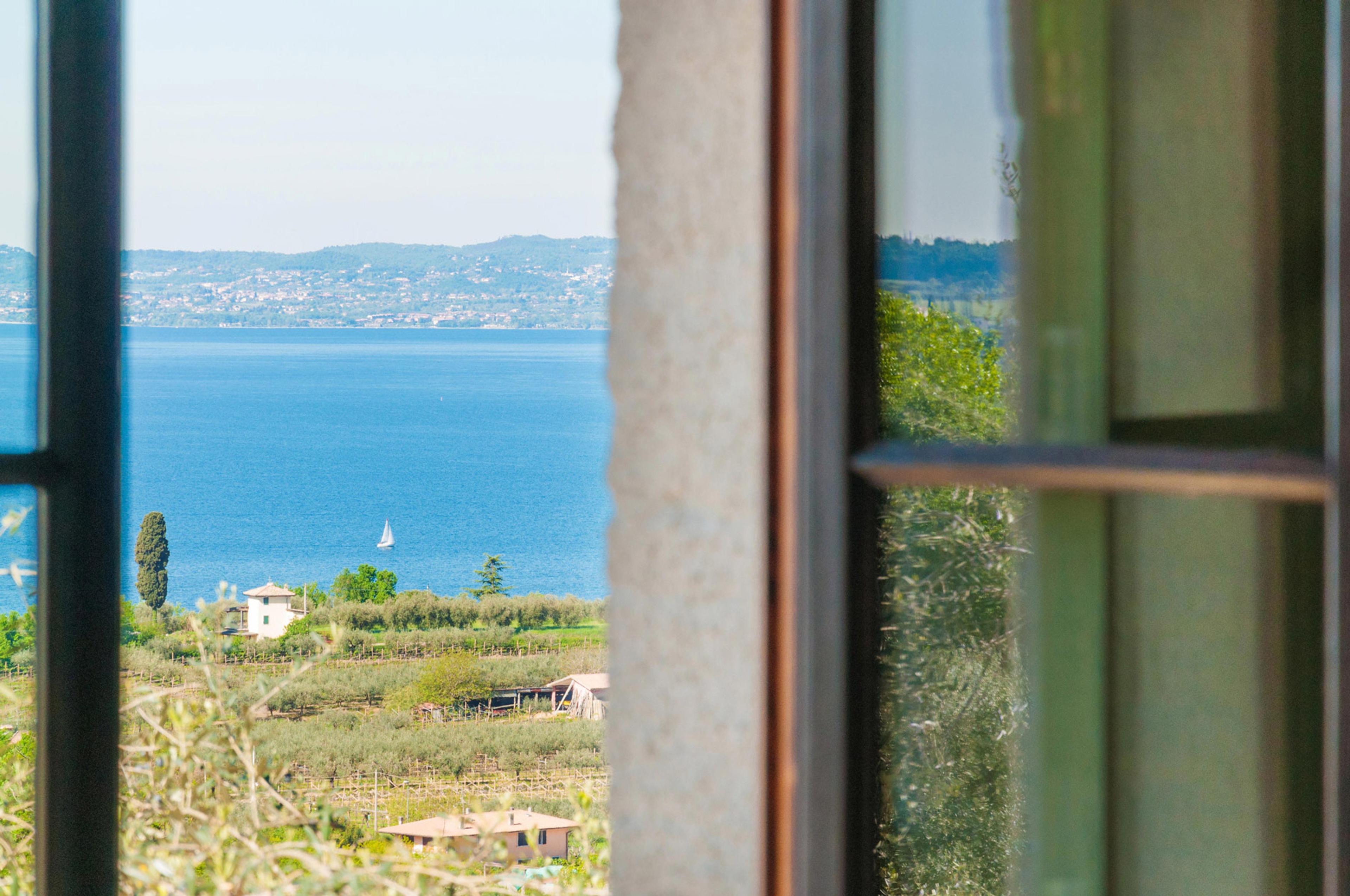 A lake view framed by an open window, with a sailboat on the water and vineyards and houses below.