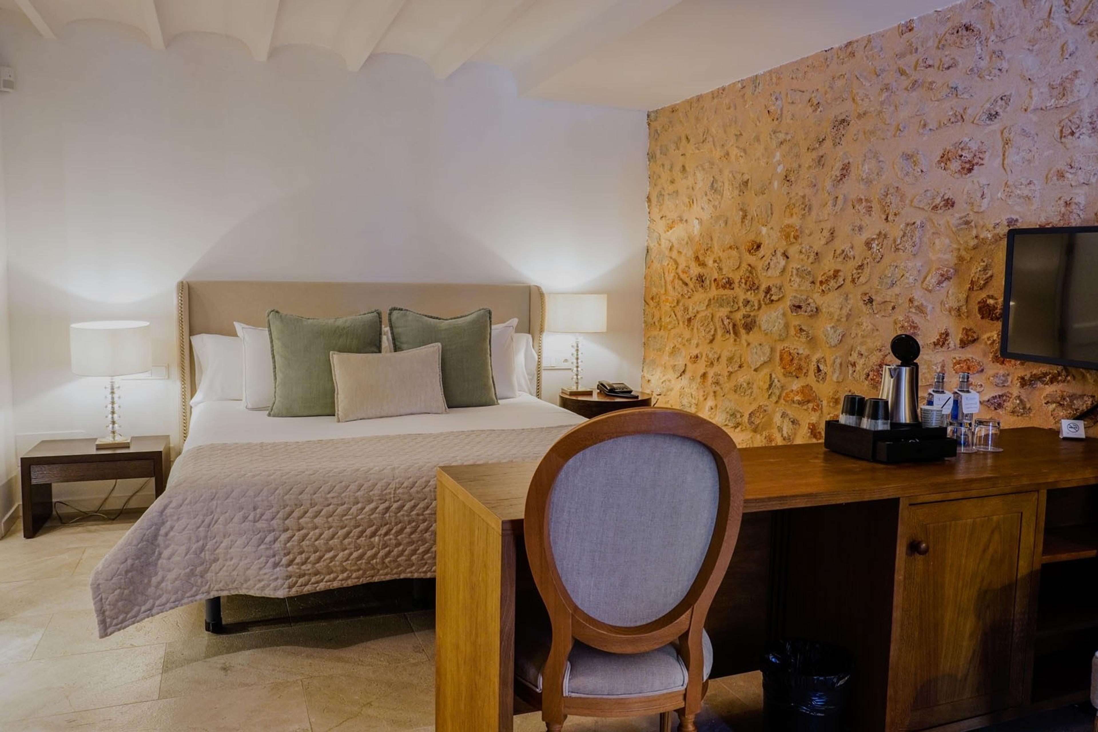 A hotel room with a bed dressed in green and taupe pillows, a stone accent wall, a wooden desk and chair, a tea tray, and a mounted television under a beamed ceiling.