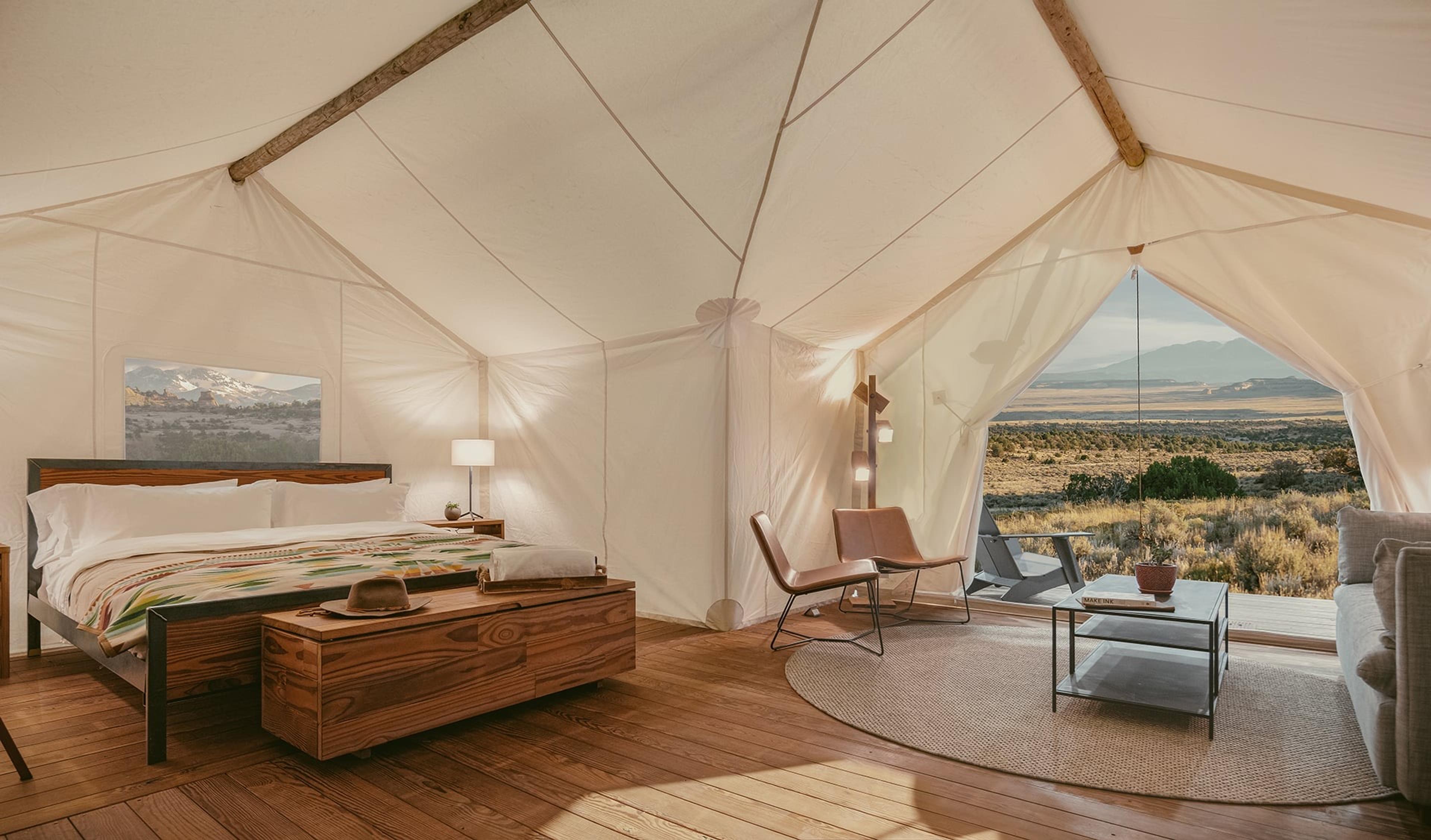 A bedroom inside a canvas tent, with a king bed under a striped blanket, a wooden bench at its foot, two leather chairs on a round rug, and the tent door open on a view of scrubland and mountains.