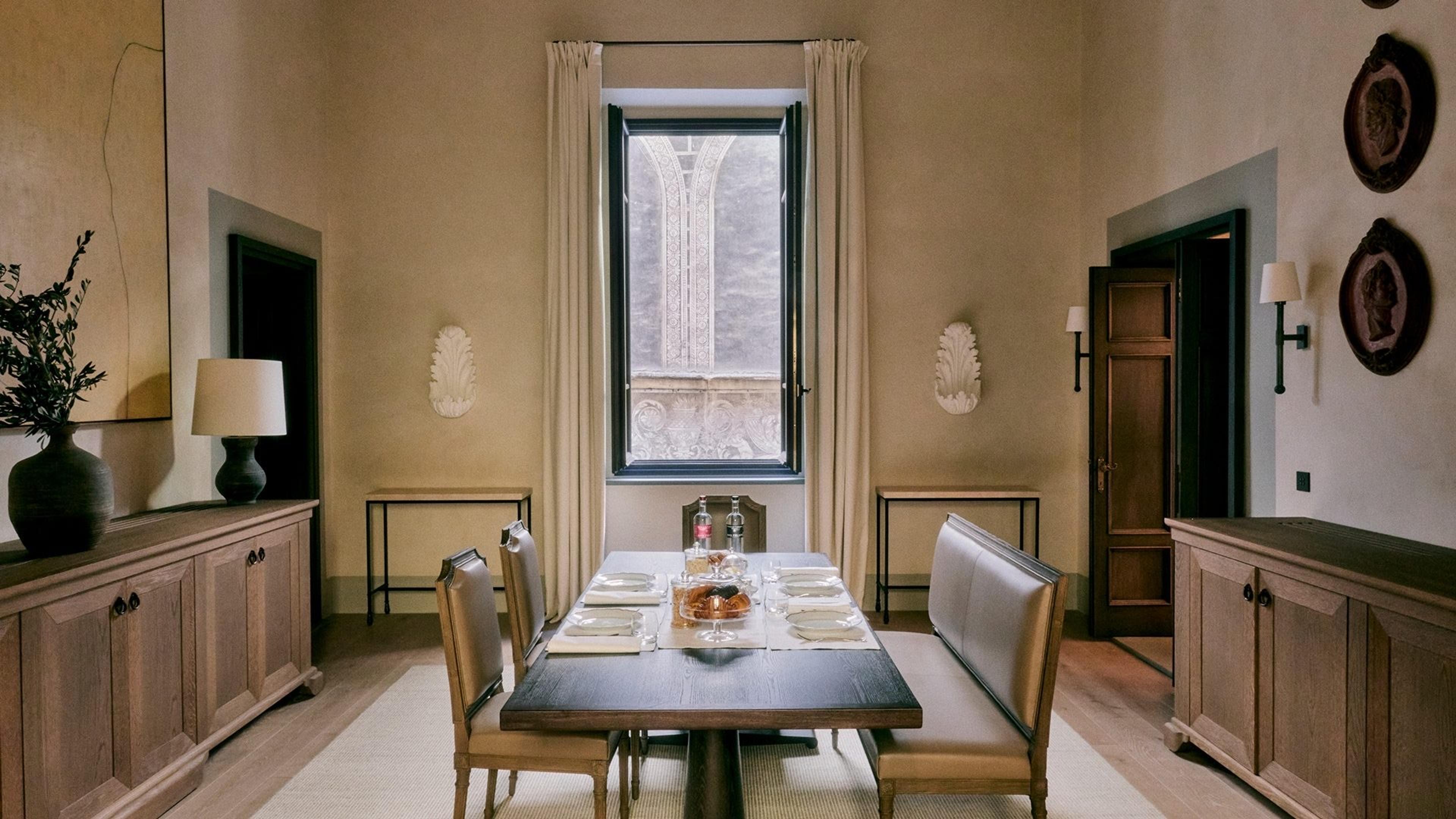A dining table laid with plates, glasses and a cheese board beneath a tall arched window showing carved stone tracery outside.