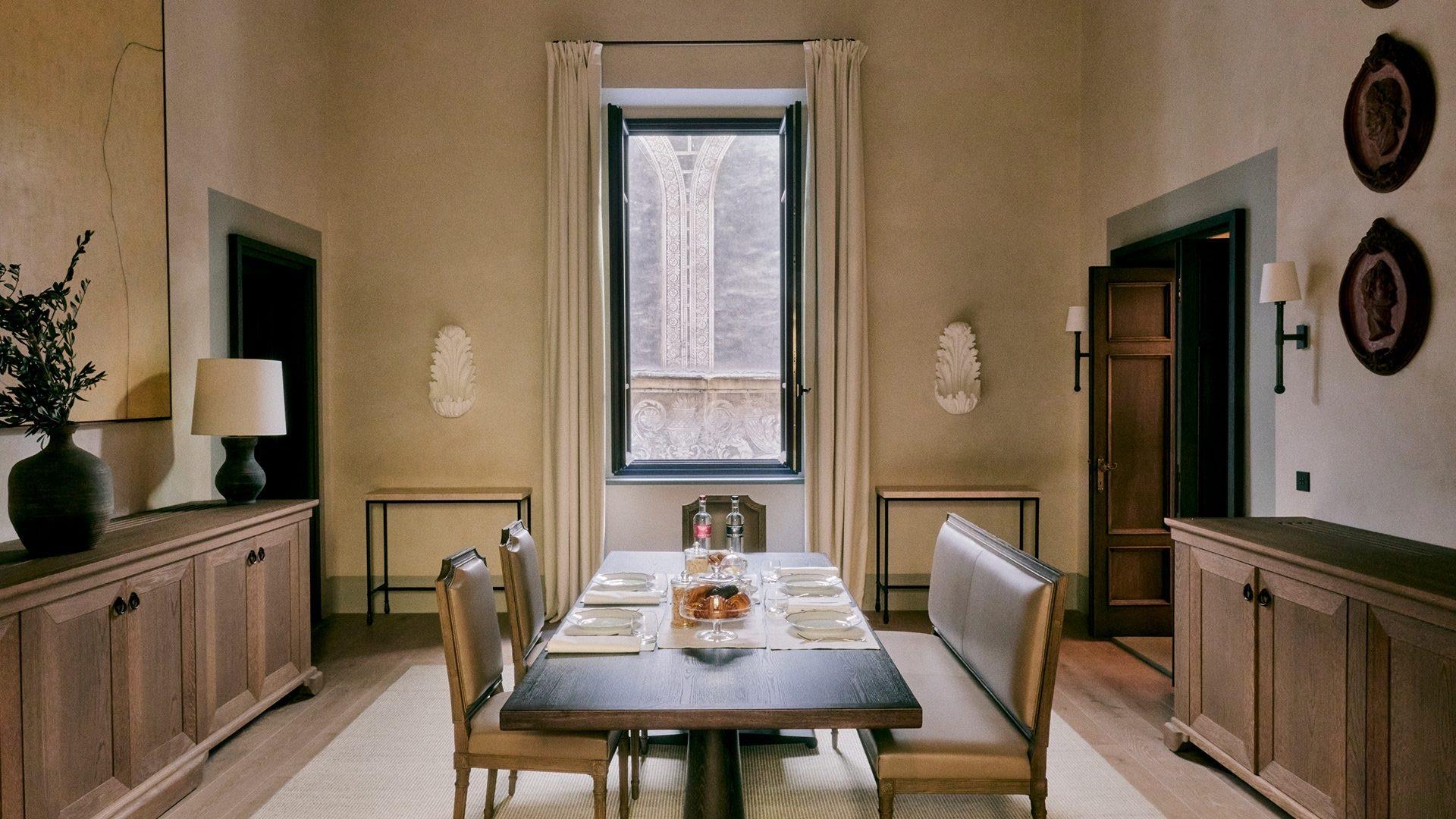 A dining table laid with plates, glasses and a cheese board beneath a tall arched window showing carved stone tracery outside.