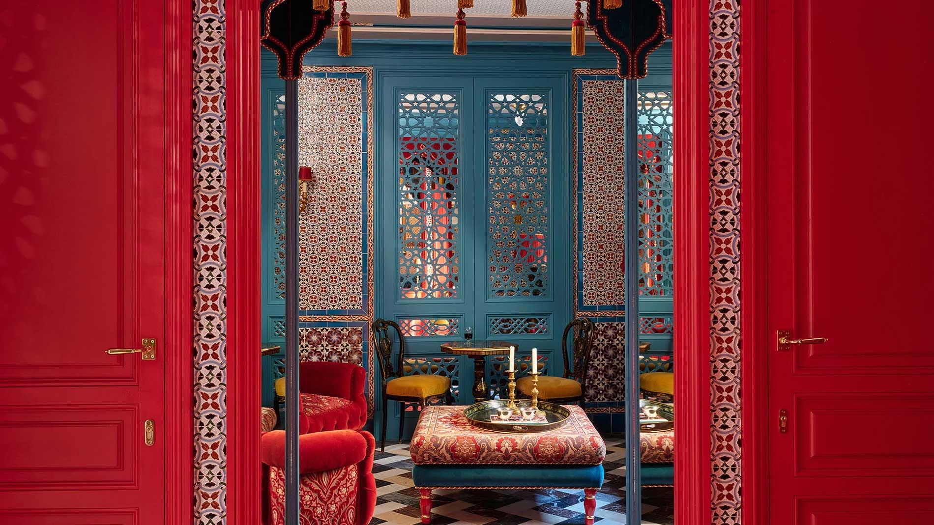 An open red double door revealing a small sitting room with carved blue screens, a patterned ottoman, gold chairs and tasselled lamps.