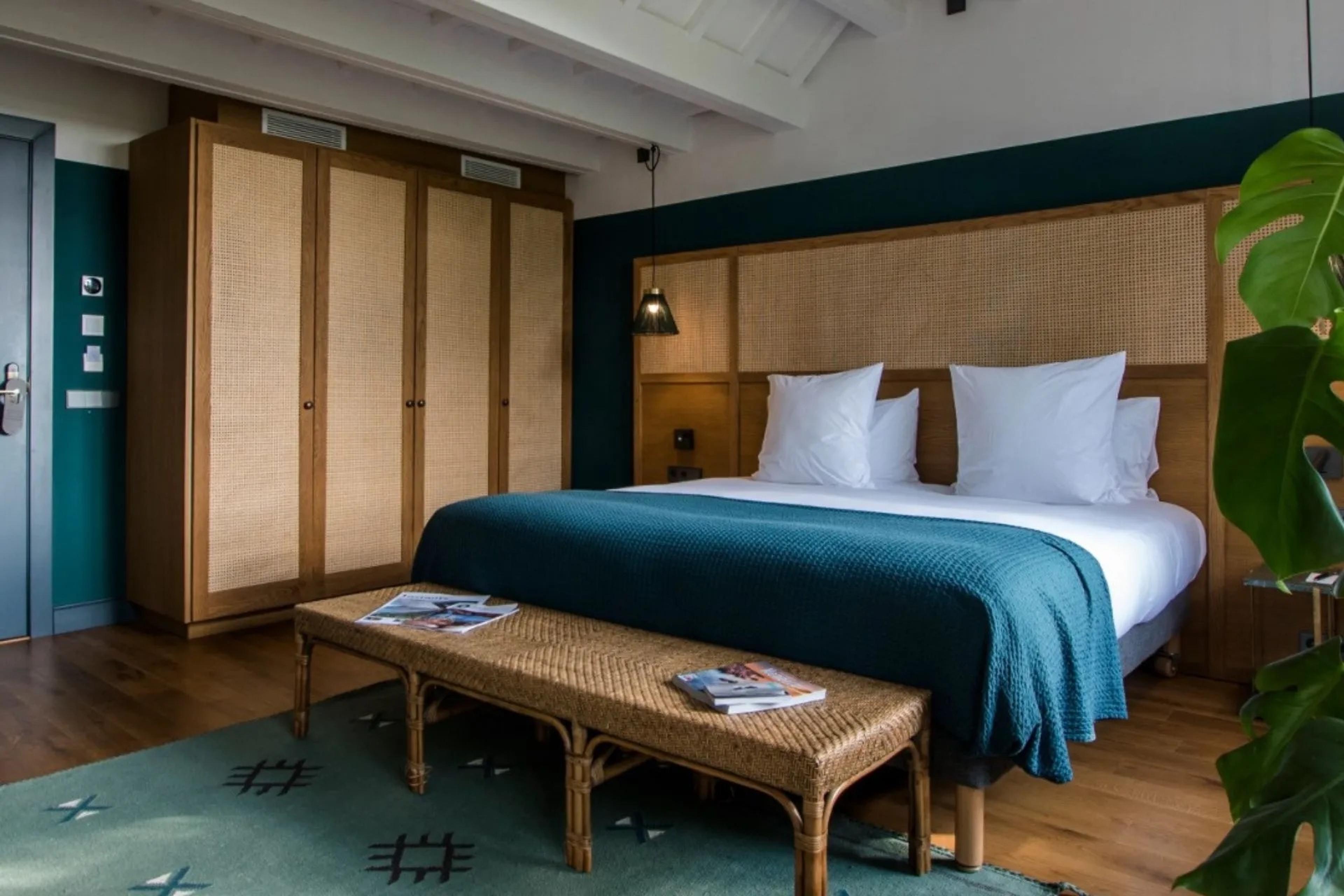 a bedroom with a woven cane headboard and wardrobe, a teal throw on the bed and a rattan bench holding magazines at its foot