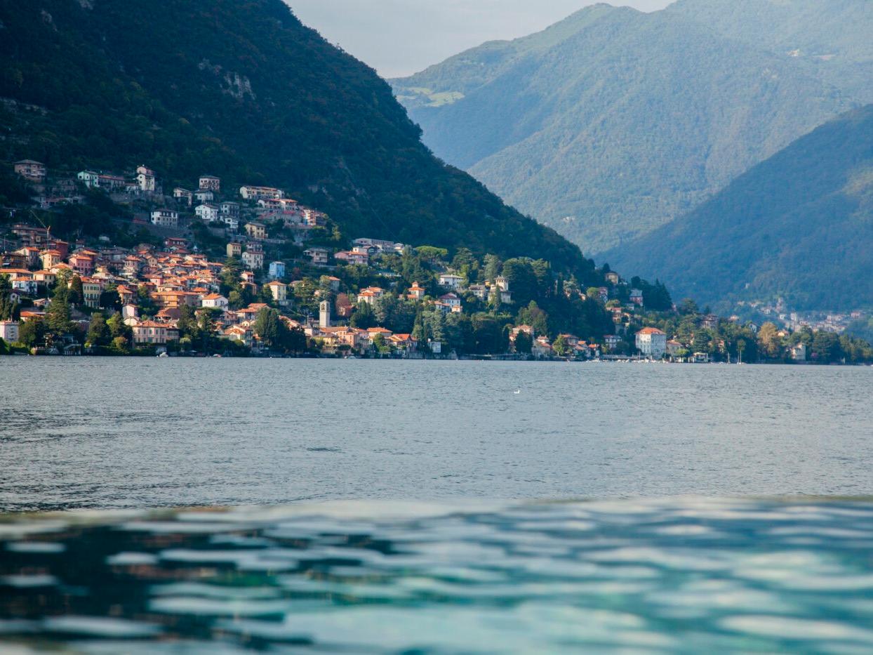 Pastel houses of a lakeside village climbing a wooded slope above Lake Como, mountains folding away behind, seen from Il Sereno at water level