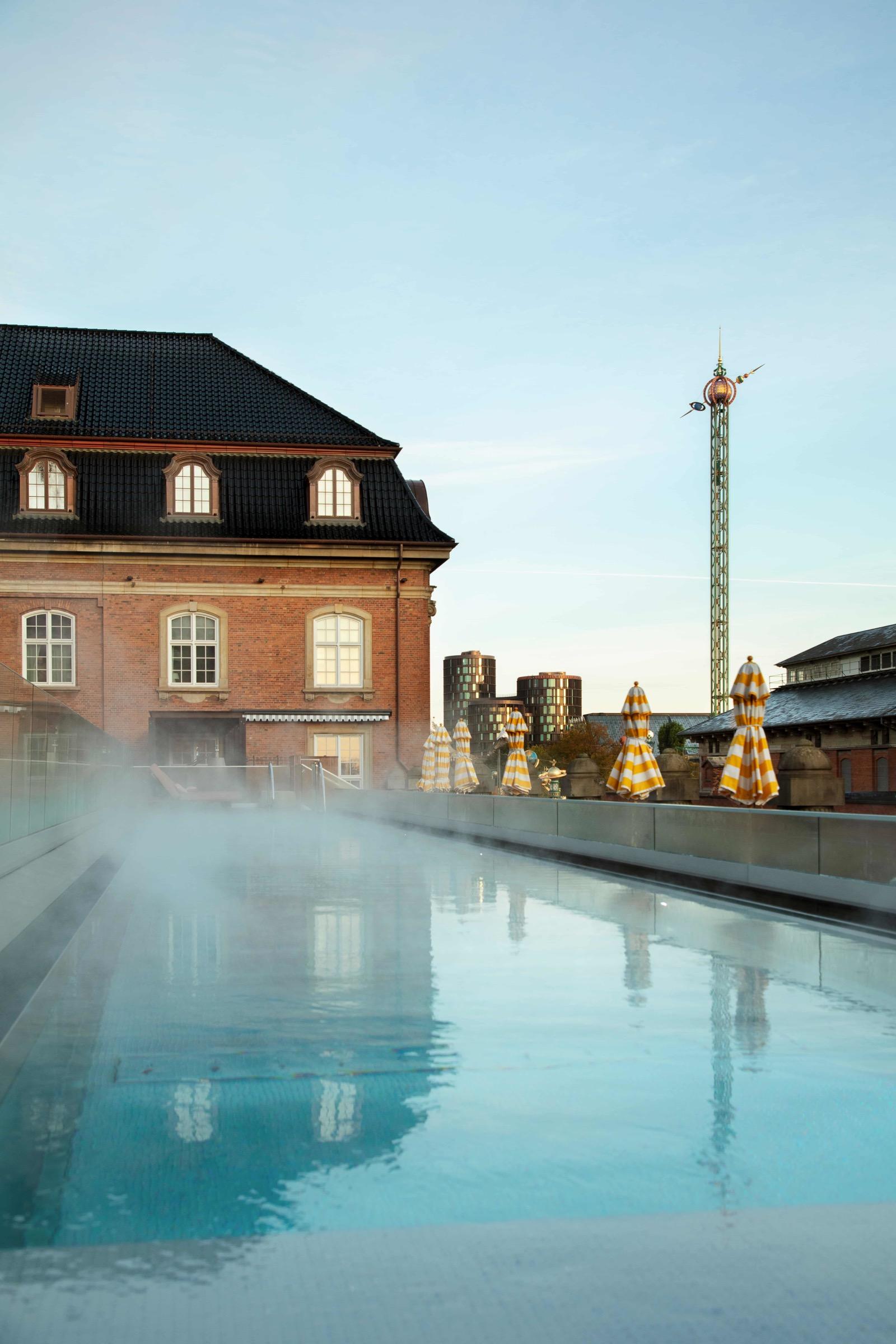 A steaming rooftop pool beside a red brick building with dormer windows, striped umbrellas and a tower visible in the distance under a pale sky.