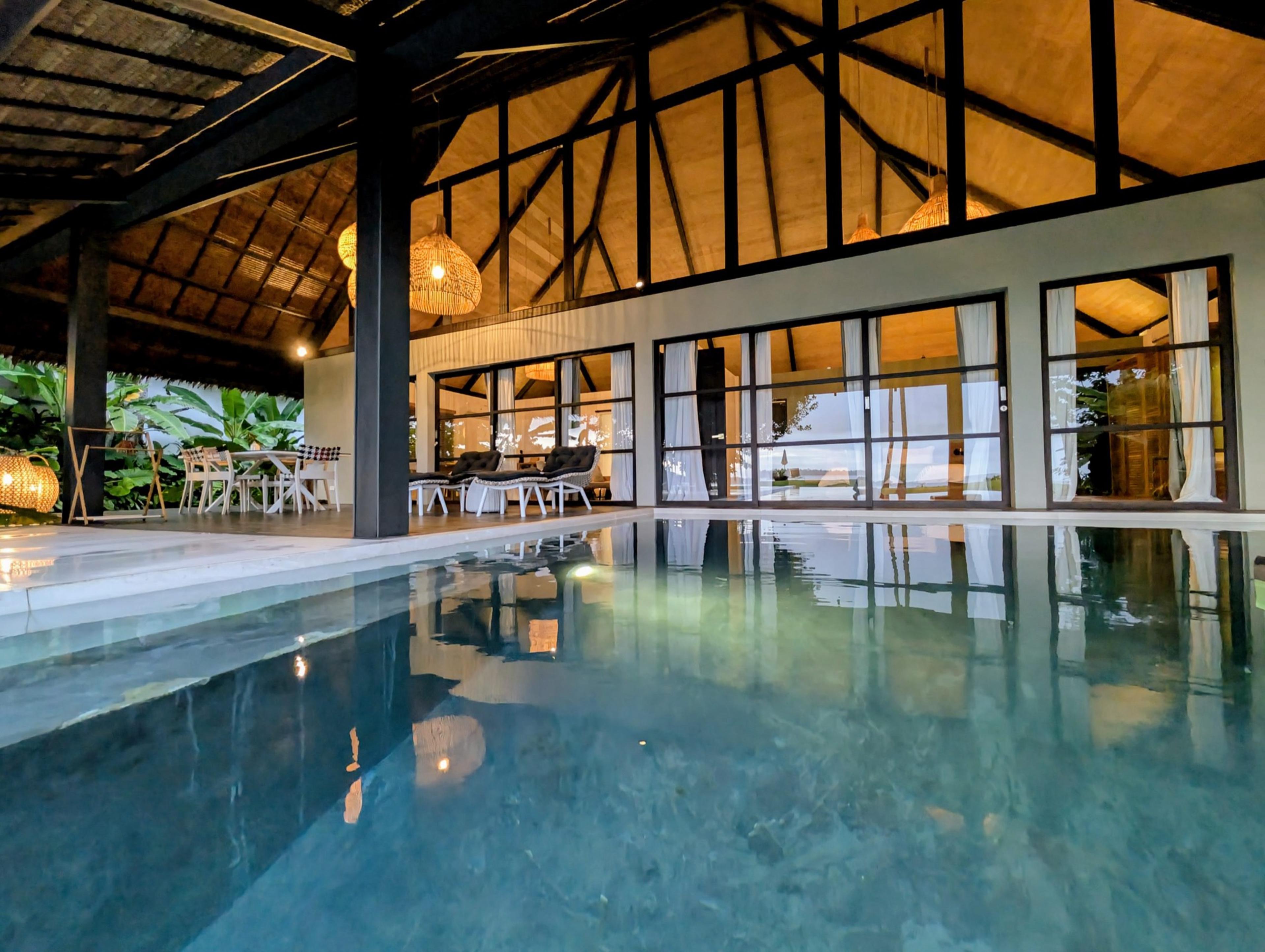 A pool lit at dusk beside an open-sided pavilion with warm light glowing through its glass doors.