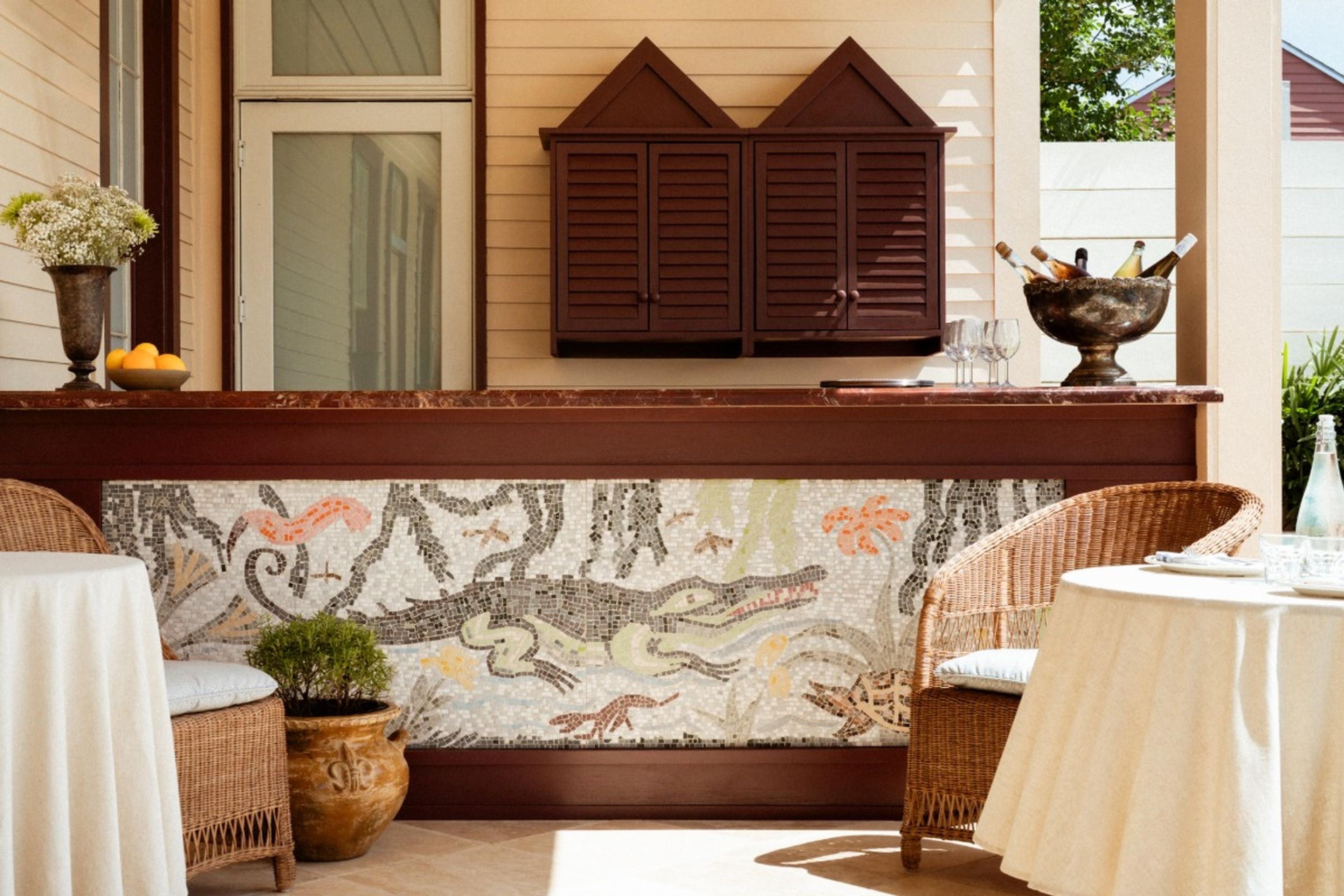 An outdoor bar with a marble counter, a mosaic mural of an alligator among plants, wicker chairs, and two round tables set for dining under a porch roof.