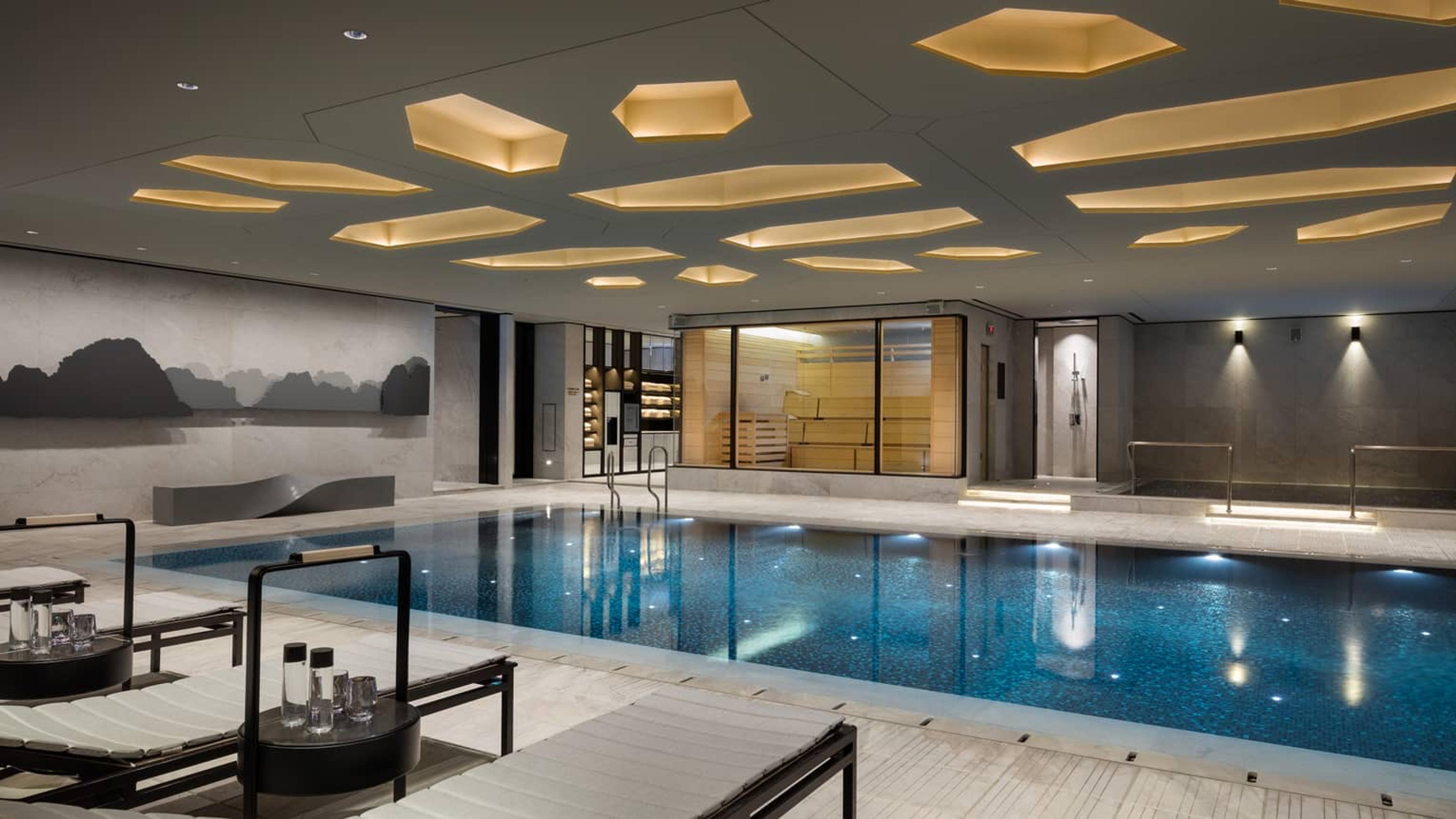 An indoor swimming pool with loungers and a drinks trolley at its edge, a glass-walled sauna, and a honeycomb-patterned lit ceiling.
