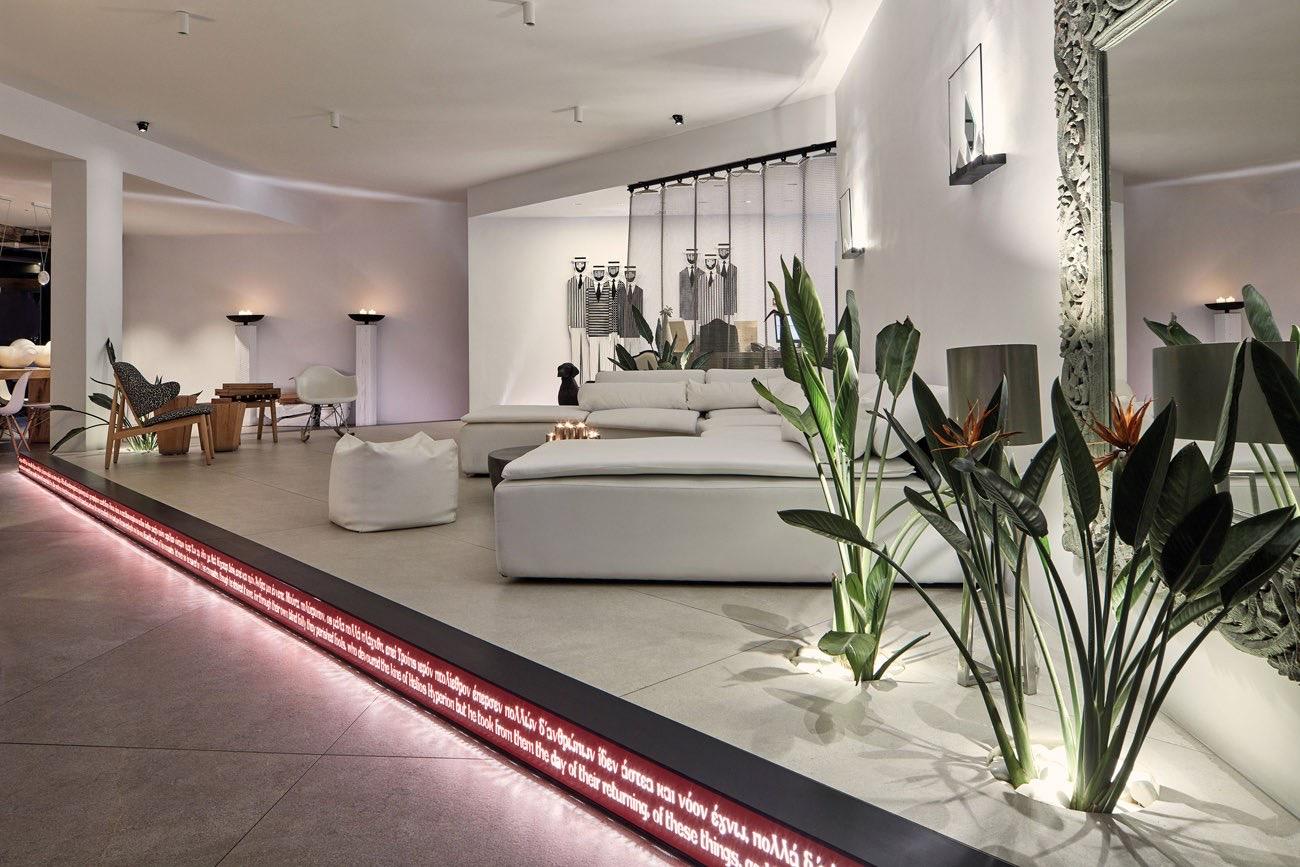 A lounge with white sofas, a bean bag, tall plants and a mirror in a carved frame, with a red-lit strip of Greek lettering running along the floor edge.