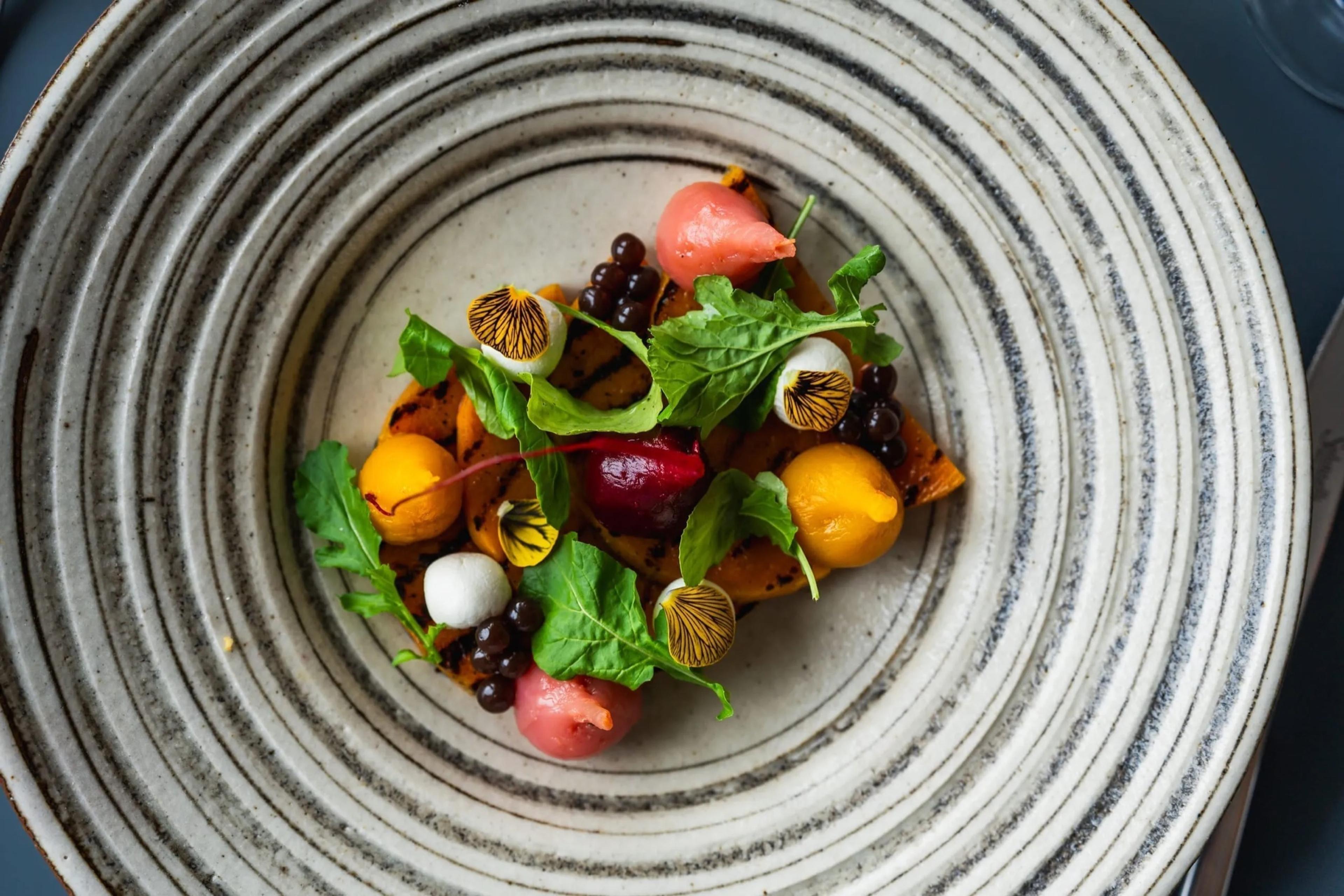 A plate of grilled orange squash topped with pink and yellow spheres, black beads, red beetroot and small leaves, seen from above on a ridged grey plate.