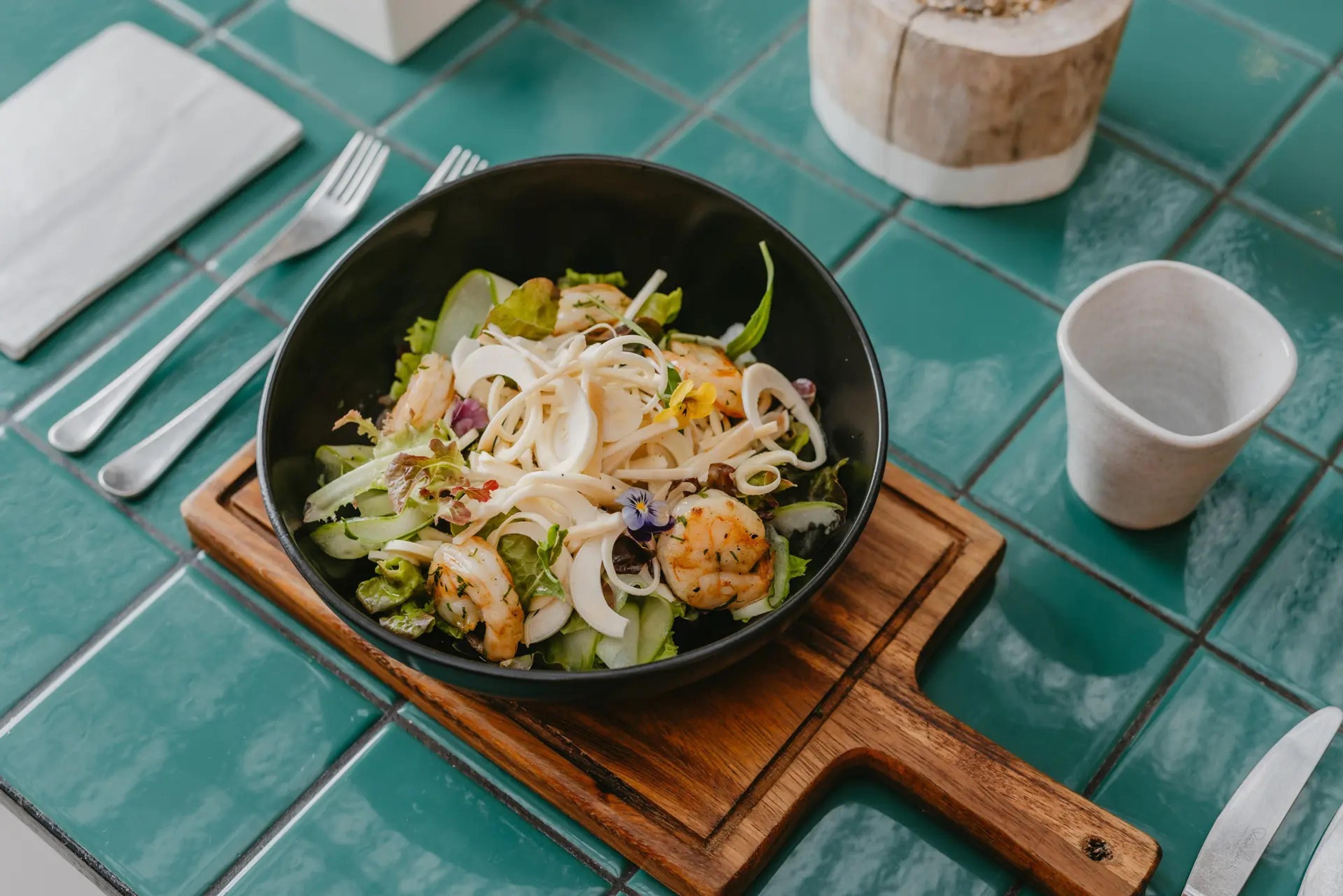 A bowl of salad with shrimp, fennel and edible flowers on a wooden board, set on a teal tiled table.