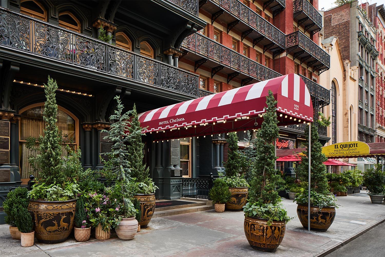 The hotel's entrance in daylight, with a red and white striped awning reading Hotel Chelsea, wrought iron balconies, and rows of potted conifers along the sidewalk.