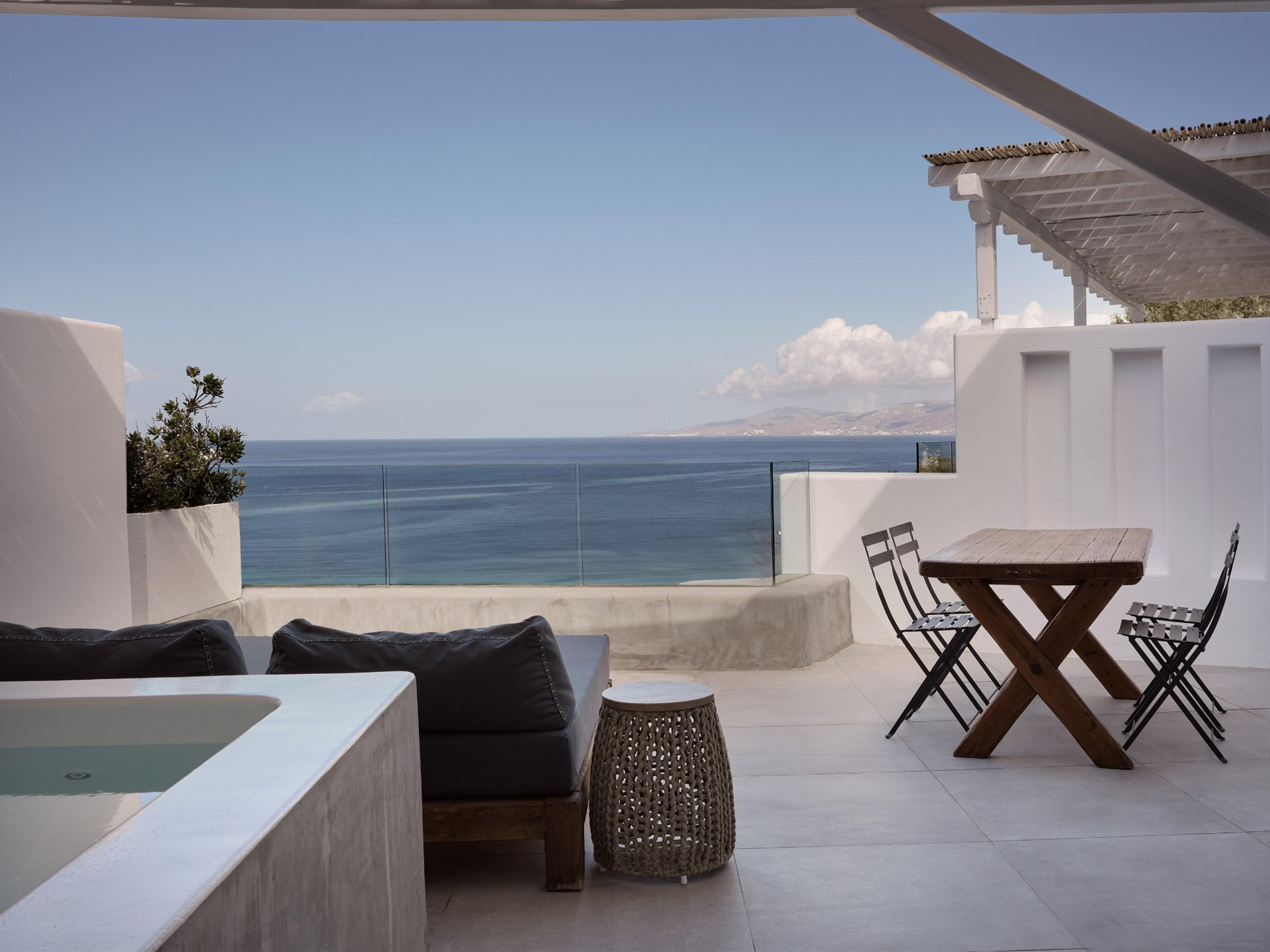 A corner terrace holds a built-in plunge pool, a dark outdoor sofa, a wooden dining table and chairs under a pergola, open to the sea on three sides.