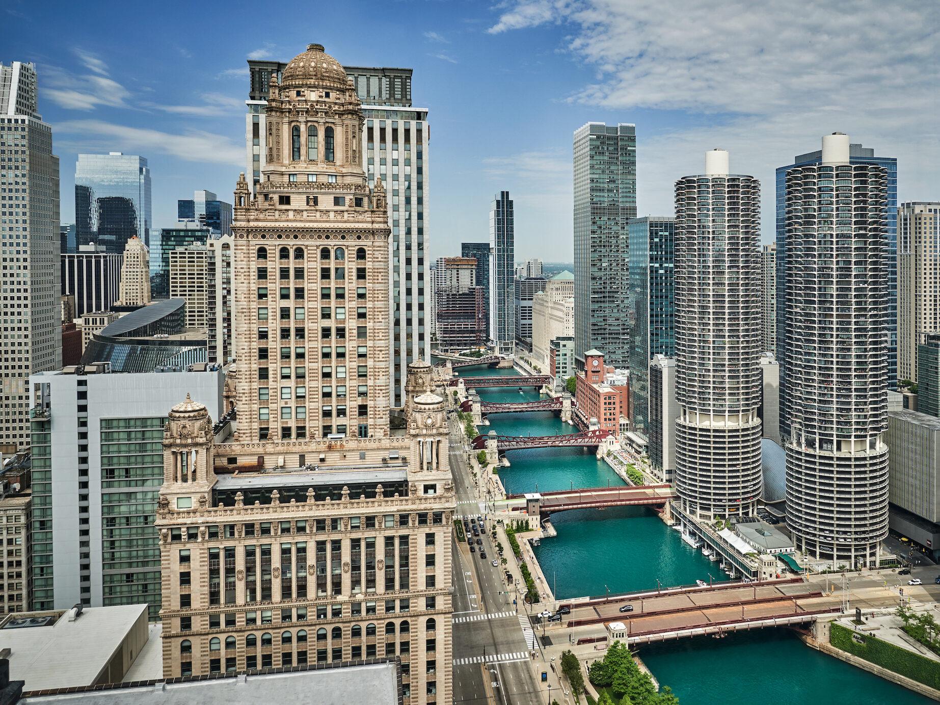 An aerial view of the Chicago River lined with bridges and skyscrapers, including a domed tower and the twin cylindrical Marina City towers.