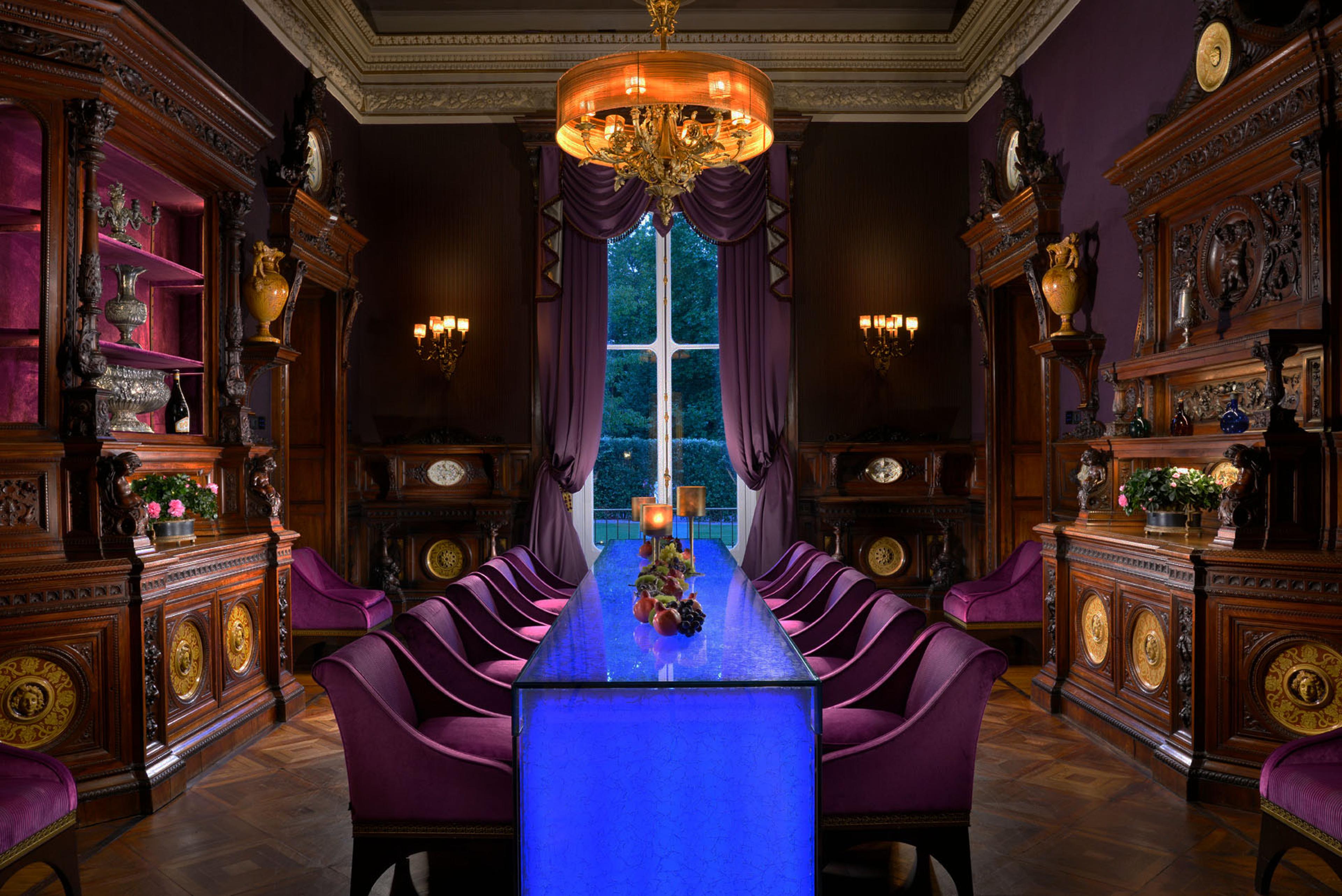 A dining room with dark carved wooden cabinets, purple chairs round a long table lit from beneath in blue, and a gilt chandelier overhead.