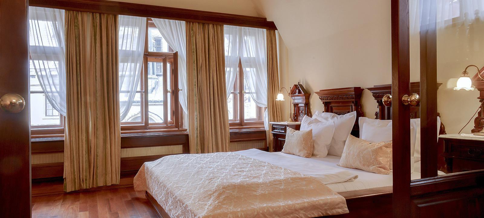 A four-poster bed with cream and gold linens in a room with tall curtained windows.
