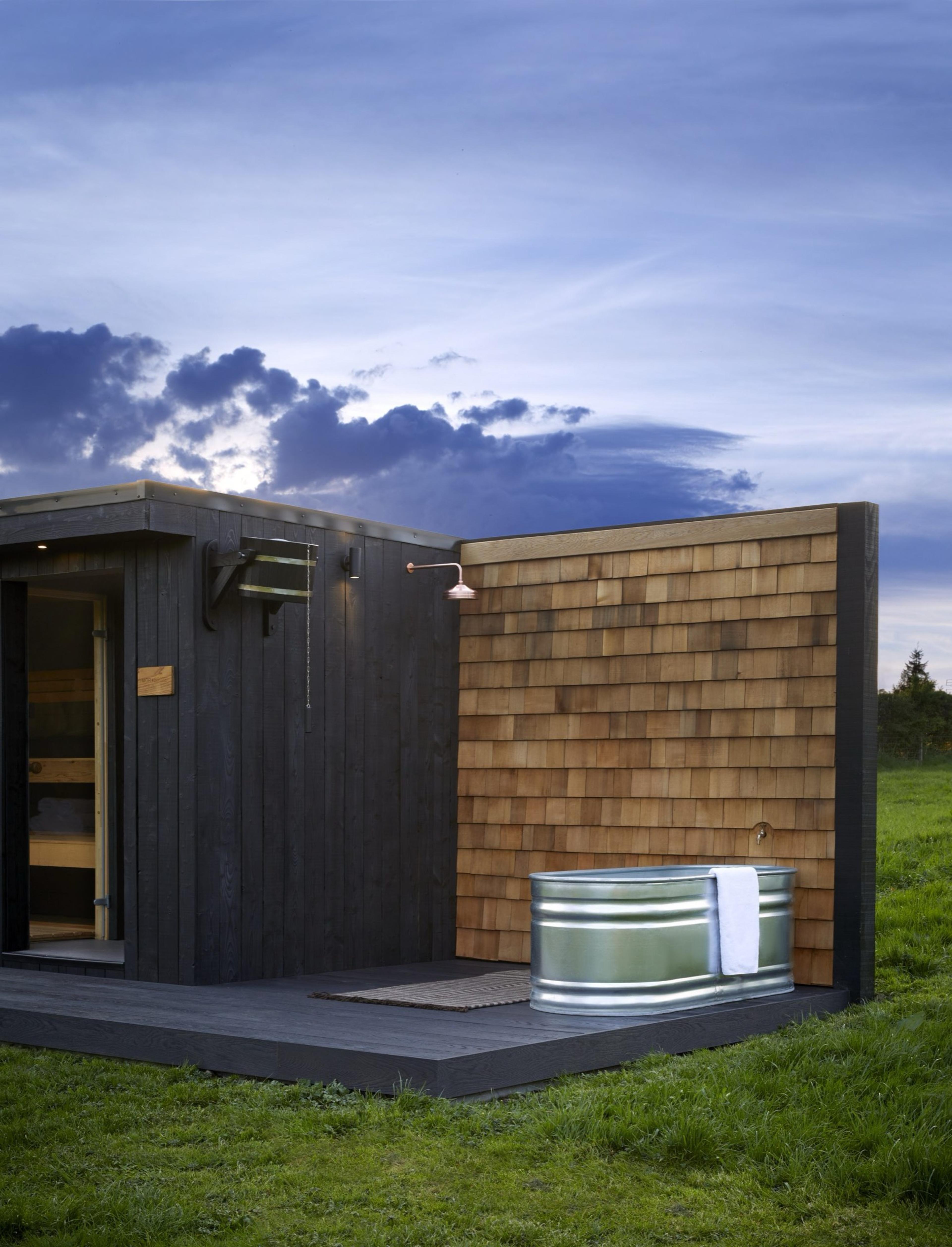 An outdoor timber cabin with a cedar shingle wall, a shower head and a round galvanised steel bath with a towel over its edge, on a deck at dusk.