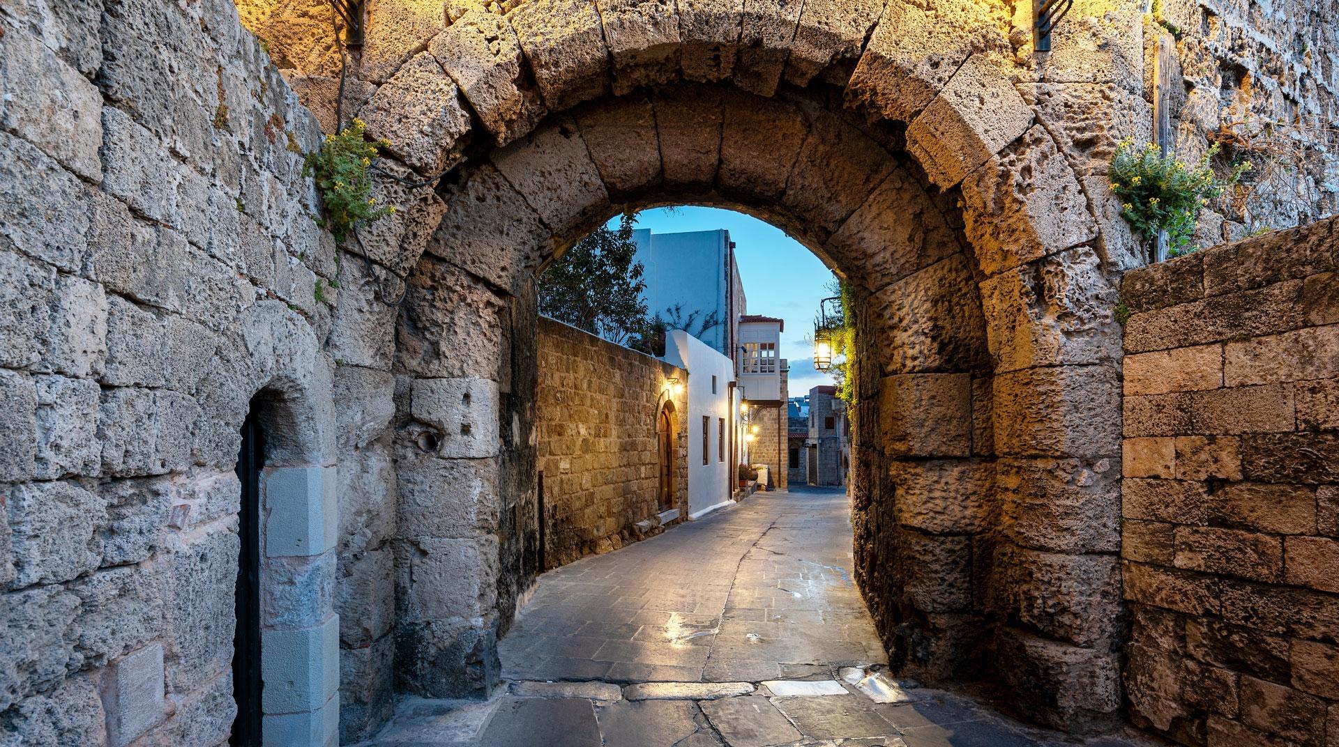 A stone archway over a cobbled street, lit at dusk with a whitewashed building visible beyond.