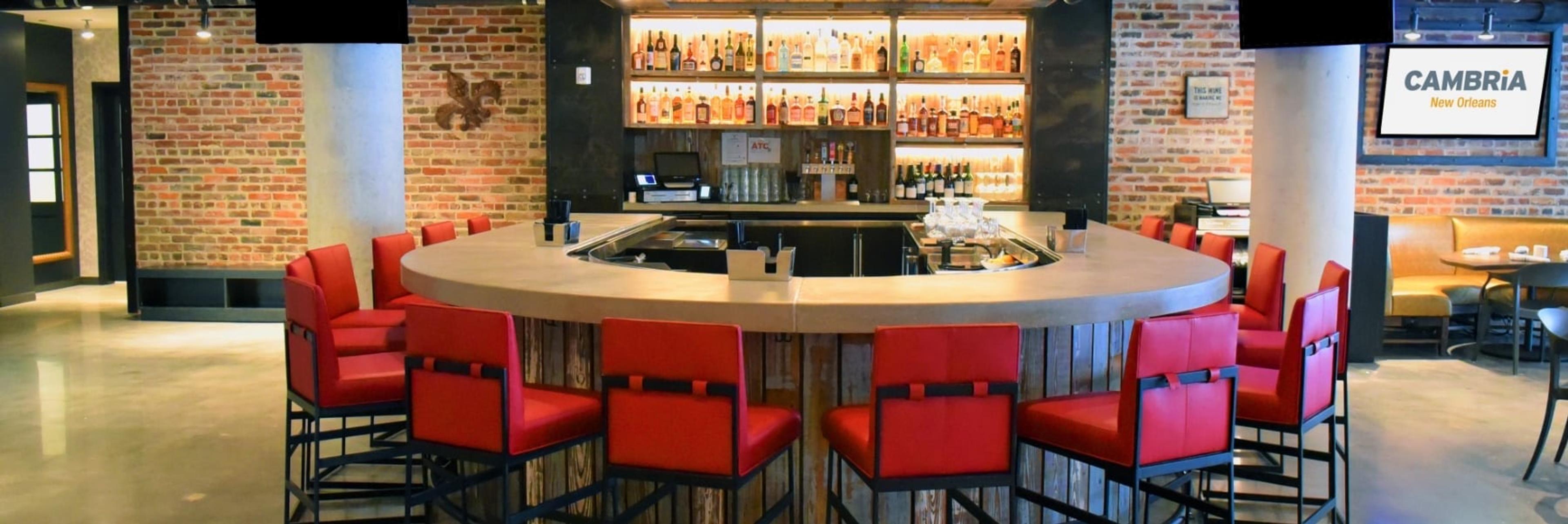 A curved hotel bar with red stools and shelves of bottles, with banquette seating and dining tables visible to one side.