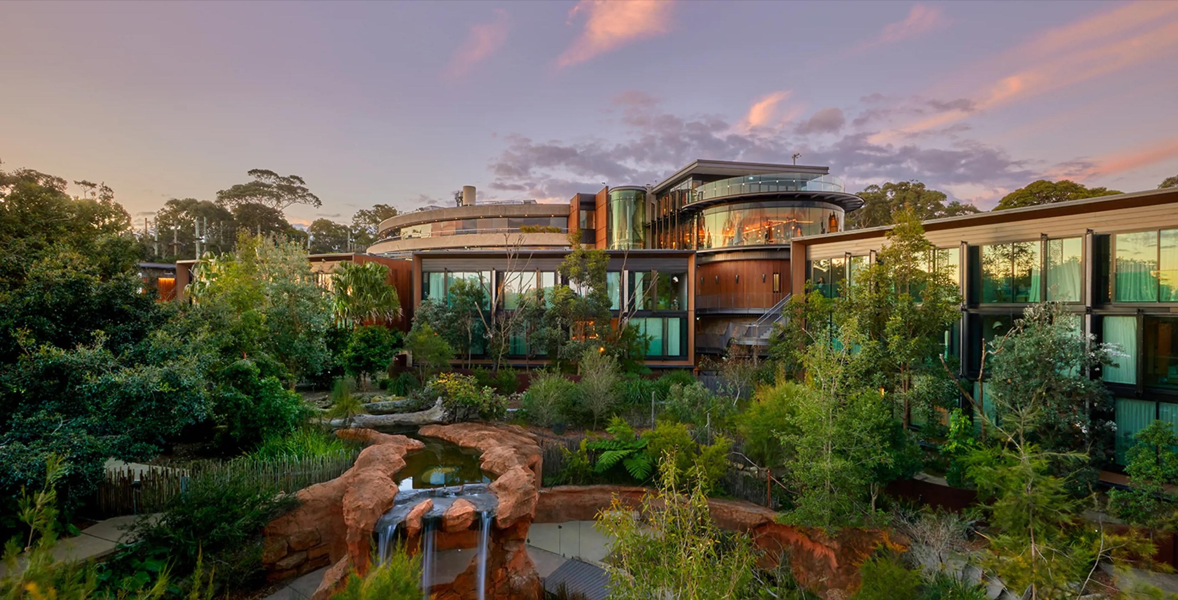 The retreat's glass and timber buildings seen from across a rock-lined water feature at dusk, surrounded by trees under a pink and orange sky.