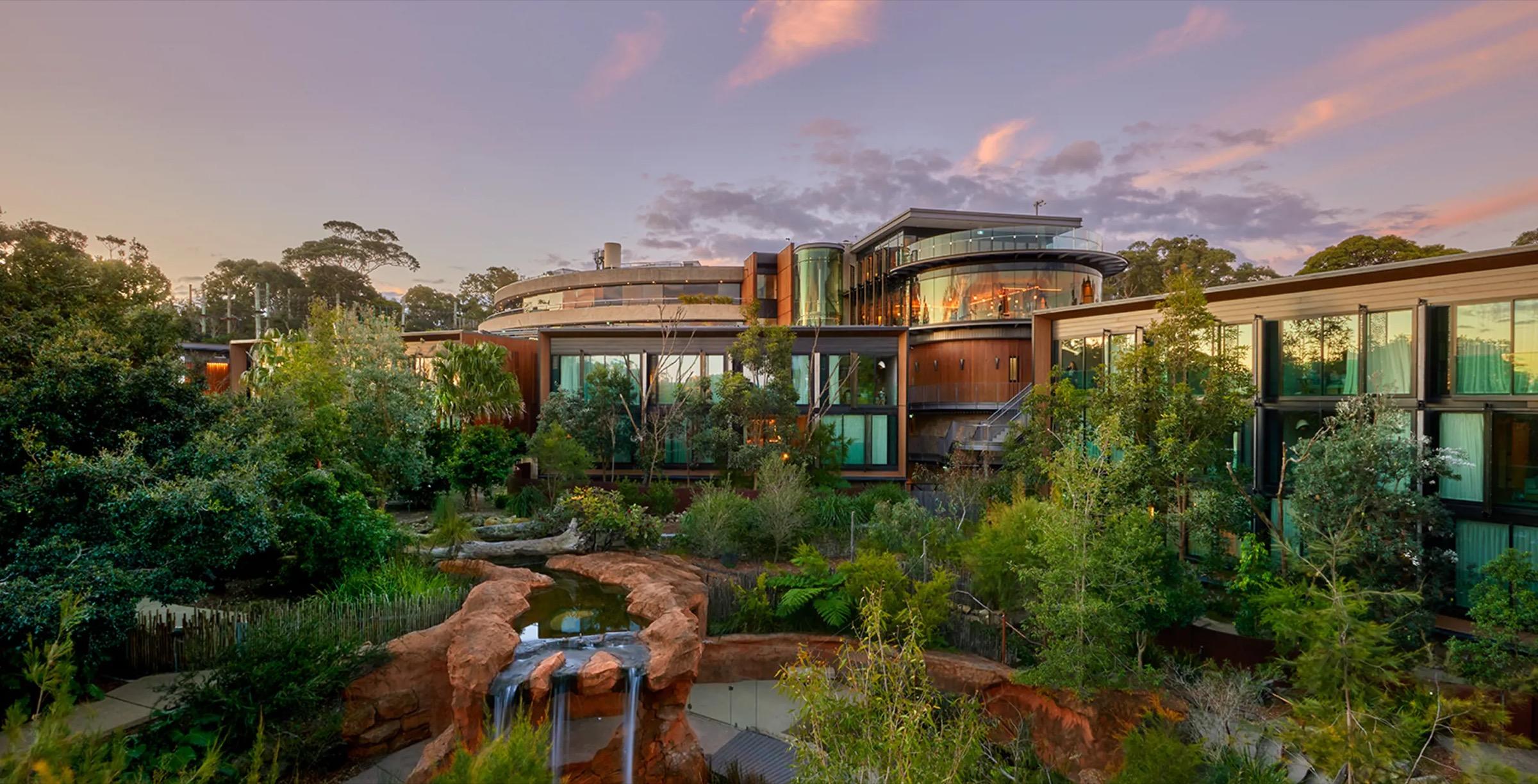The retreat's glass and timber buildings seen from across a rock-lined water feature at dusk, surrounded by trees under a pink and orange sky.