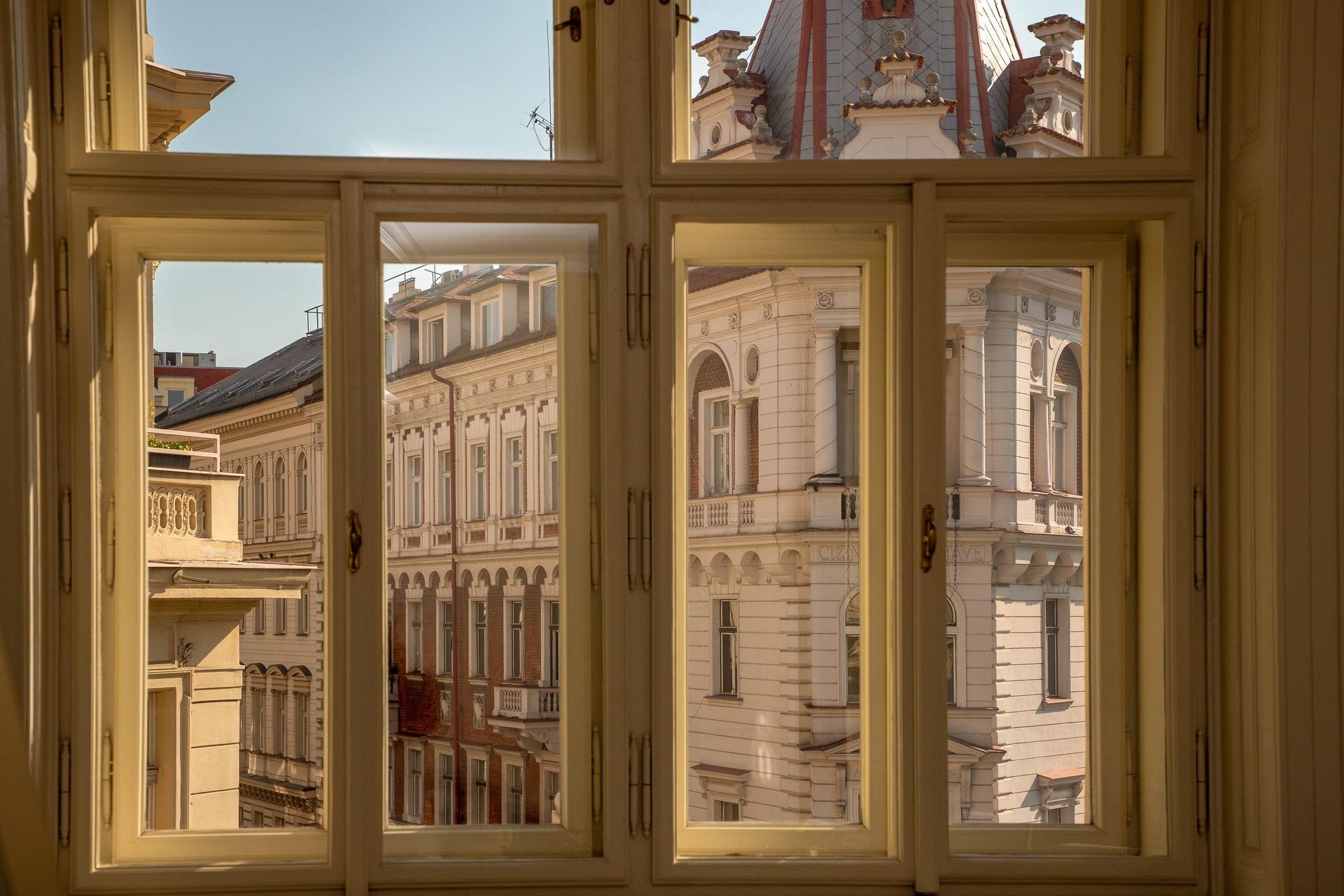 A view through an open sash window of ornate stone townhouses and a patterned roofline against a blue sky.