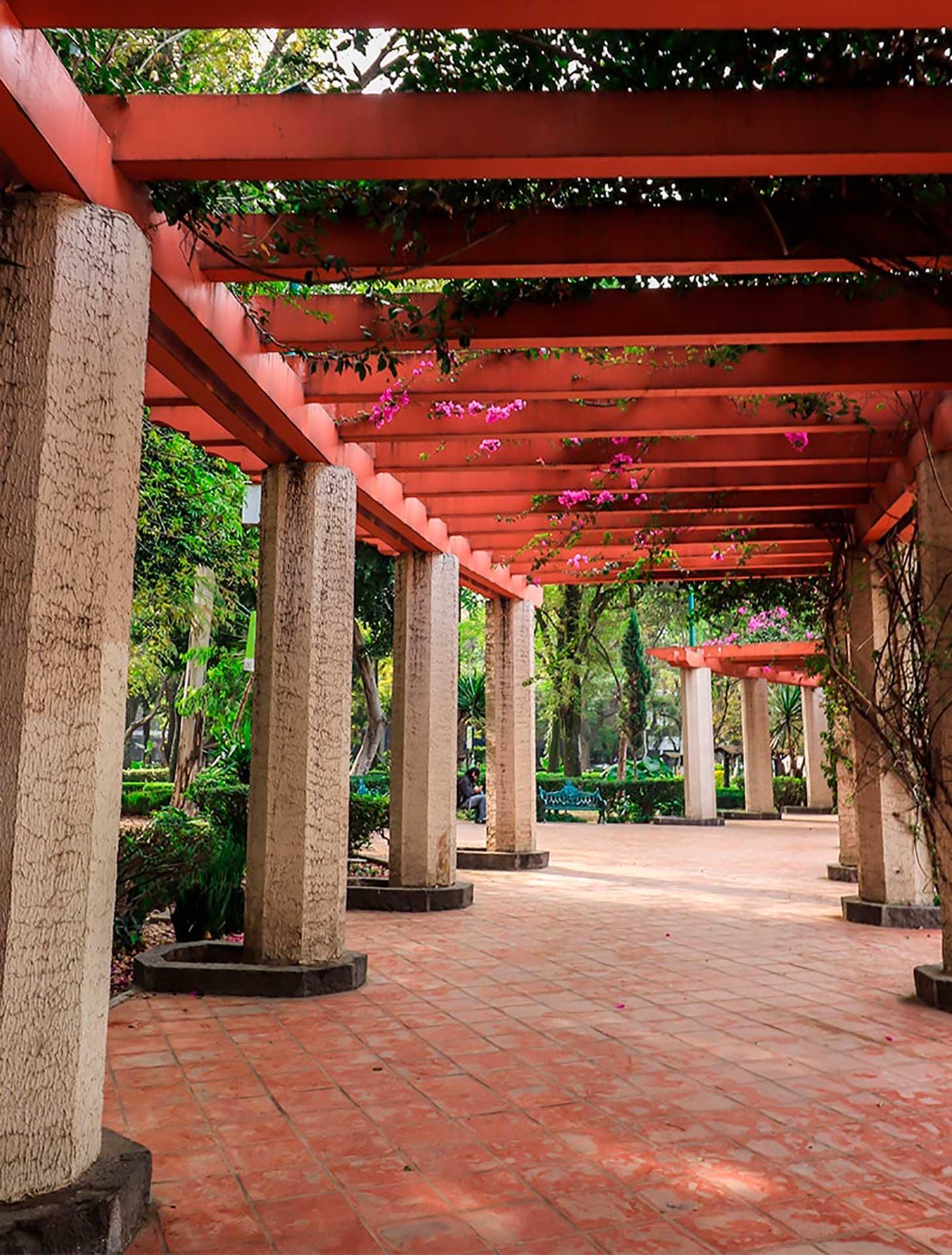 A covered walkway with round stone columns and a terracotta roof, bougainvillea blooming overhead and trees beyond, a person seated in the distance.
