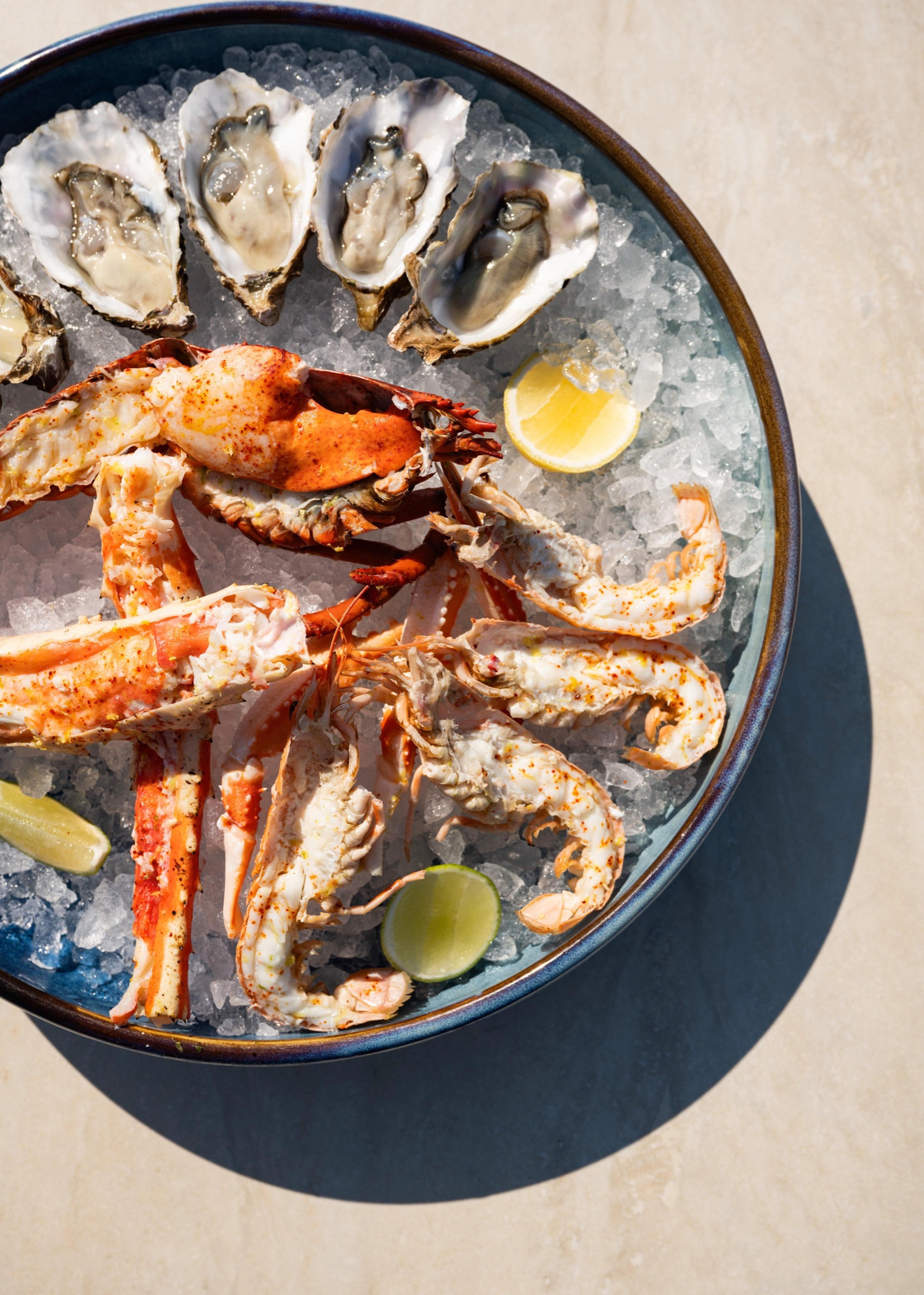 An overhead view of a round plate of oysters and lobster claws on ice, with lemon and lime wedges.