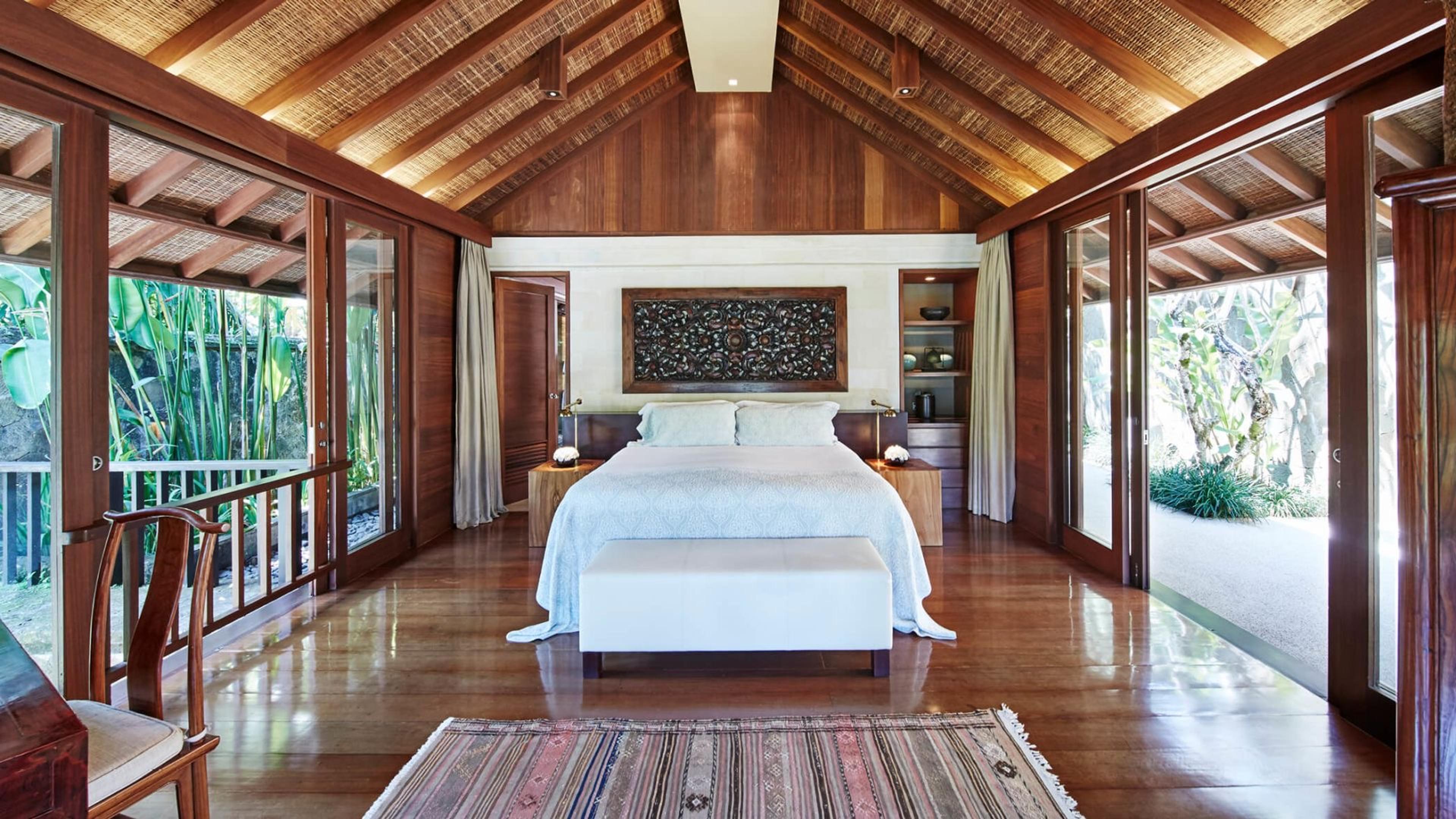 A bedroom with a vaulted thatched ceiling, a white-draped bed and a wood-carved panel above the headboard, with doors open to greenery on both sides.