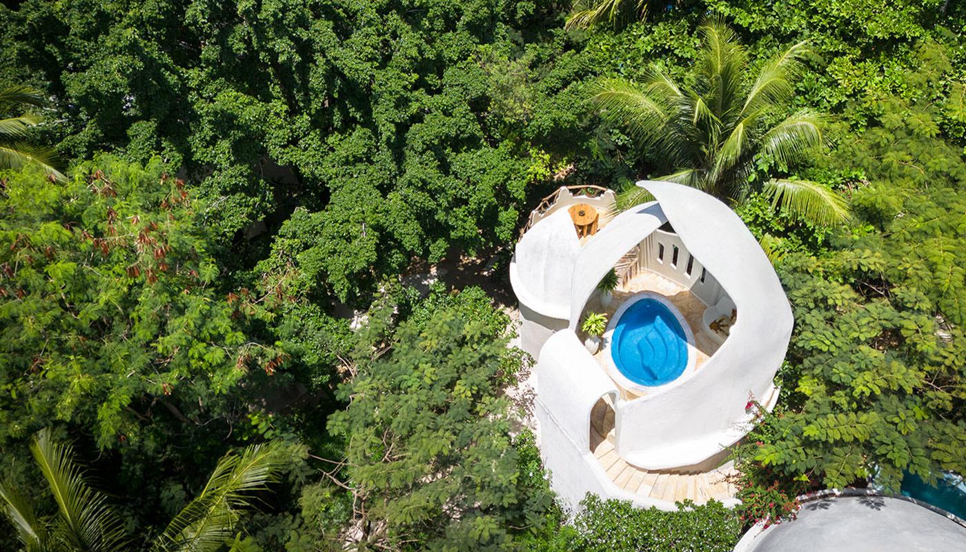 An aerial view of a round, spiral-walled villa with a plunge pool on its terrace, surrounded by dense treetops.