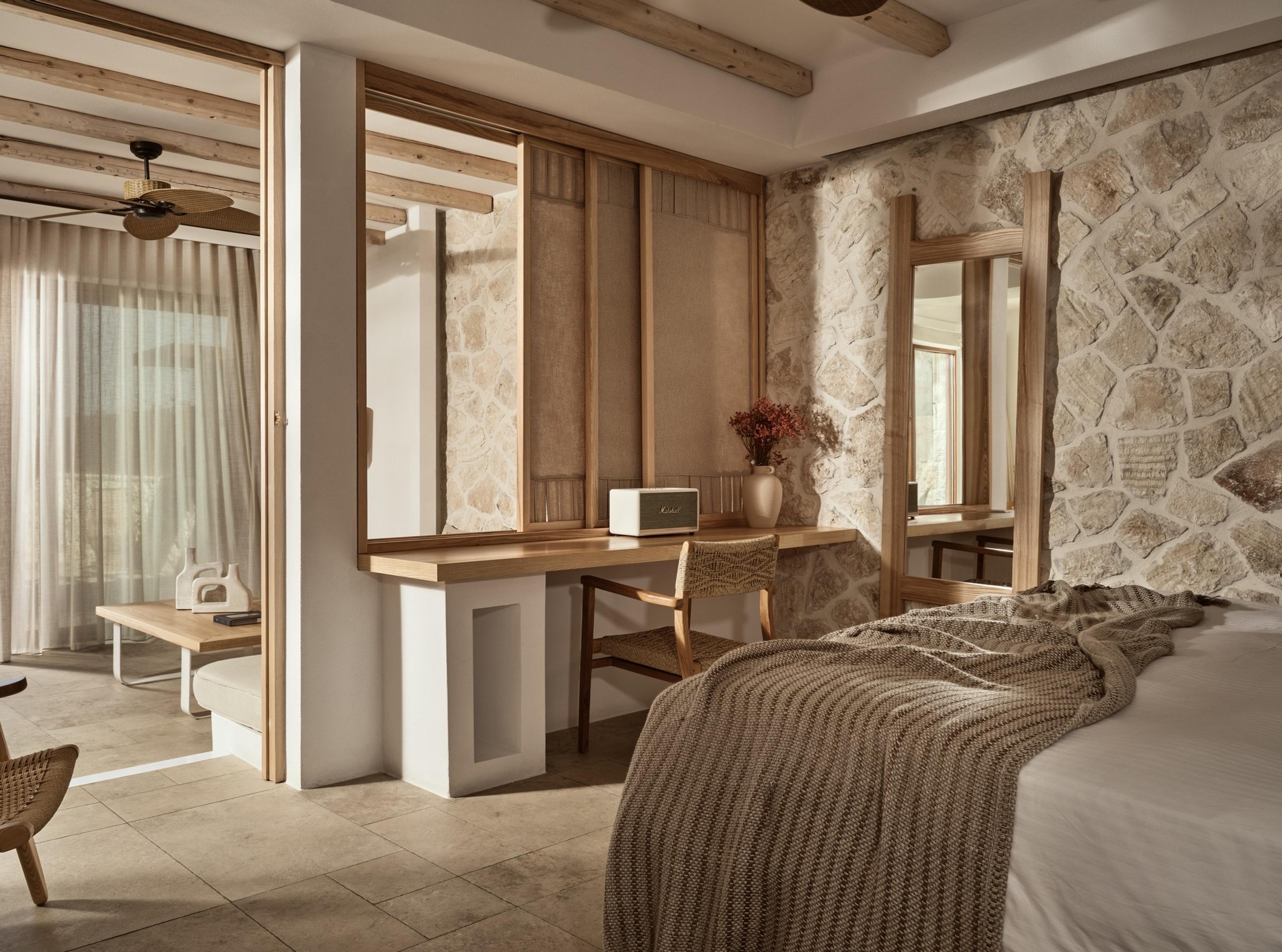 A bedroom with a stone feature wall, a wood-beamed ceiling, a woven-headboard bed and a desk below a round mirror.
