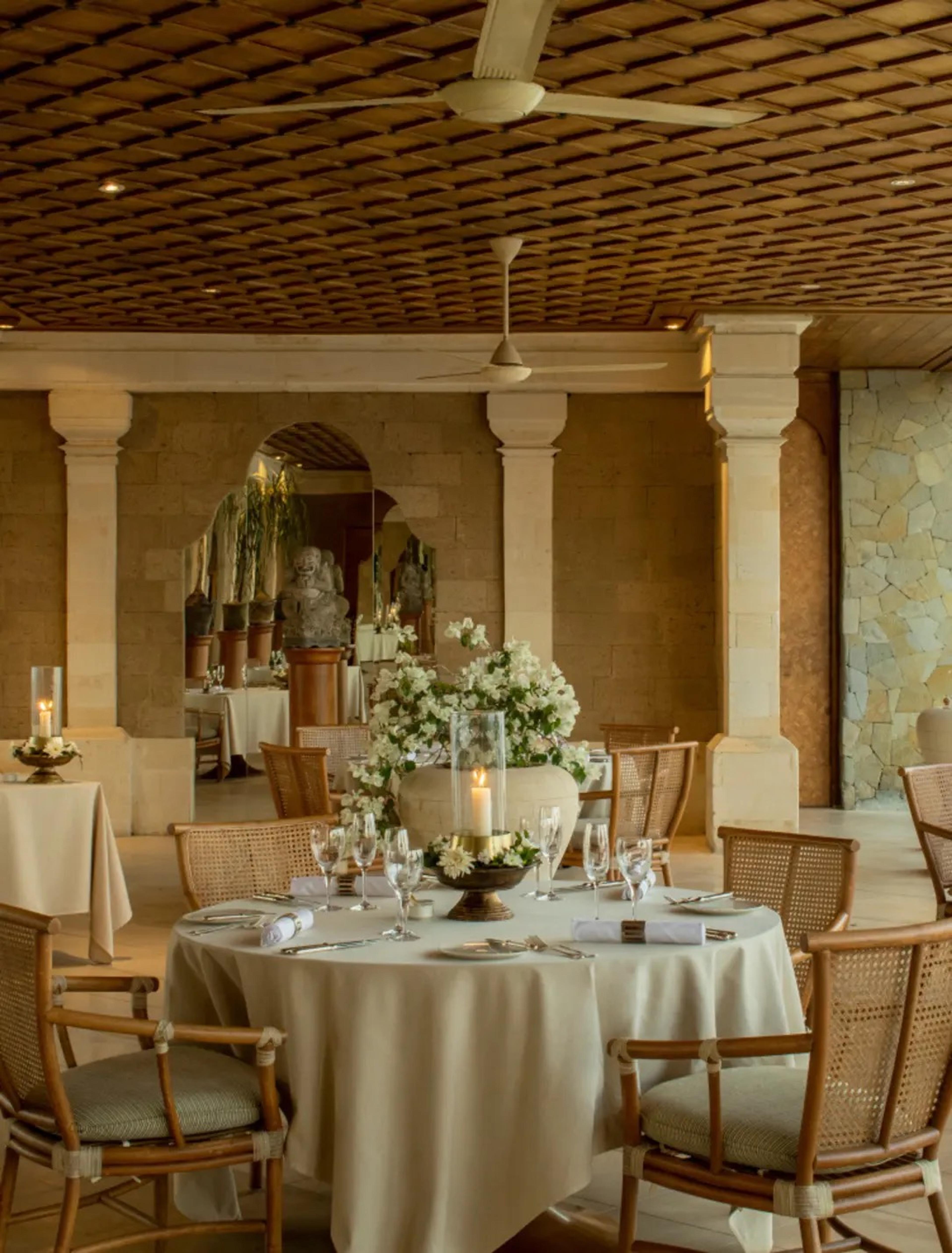 A round dining table set with glasses, candles and white flowers, inside a restaurant with stone columns and ceiling fans.