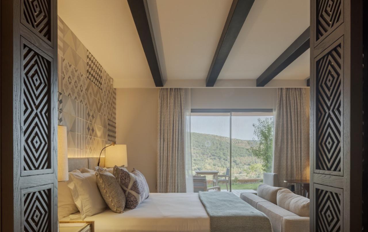 A bedroom seen through open carved wooden doors, with a bed, a patterned tile headboard, and a sliding glass door onto hills beyond.