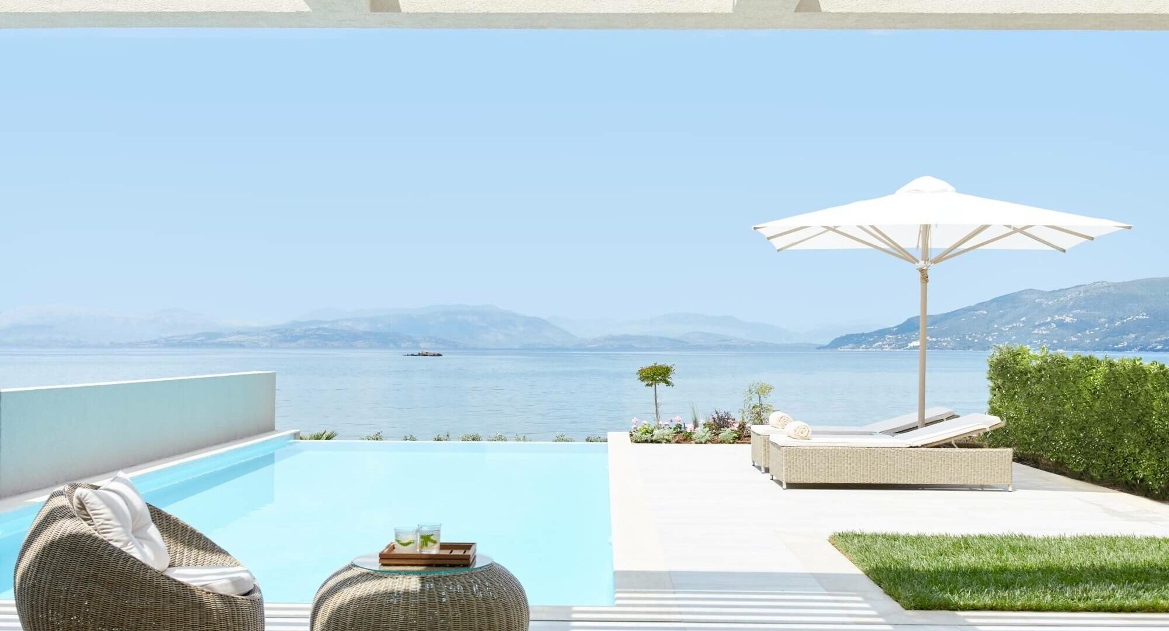 A private pool terrace with woven armchairs and a footstool in the foreground, an open umbrella and sunbeds beyond, overlooking the sea and hills.