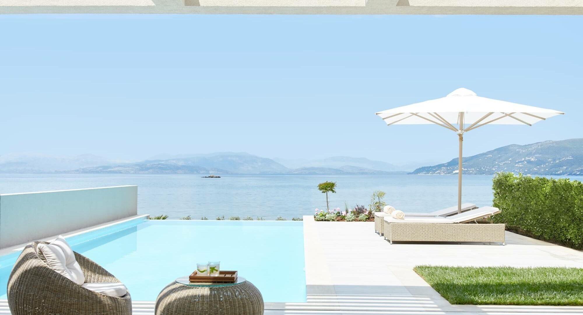 A private pool terrace with woven armchairs and a footstool in the foreground, an open umbrella and sunbeds beyond, overlooking the sea and hills.