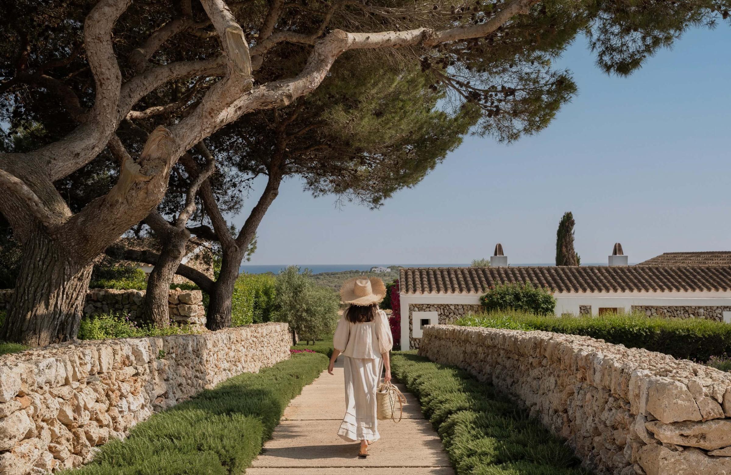 A woman in a straw hat and white dress walks away along a stone-walled path lined with pine trees, with farm buildings and the sea ahead.