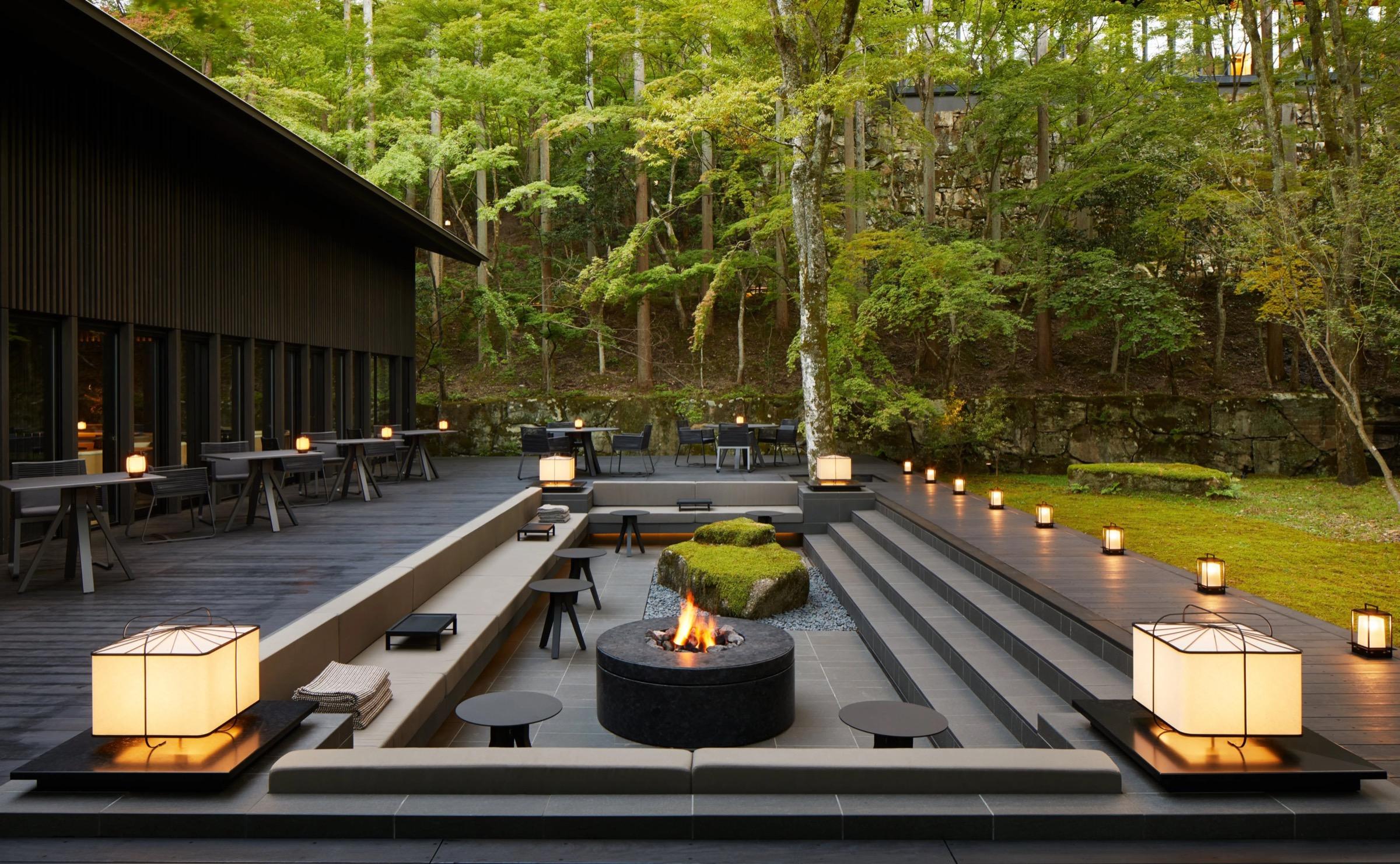 A terraced outdoor lounge at dusk, with built-in bench seating, a lit firepit, lanterns on stone steps and a dark timber building to one side, forest behind.